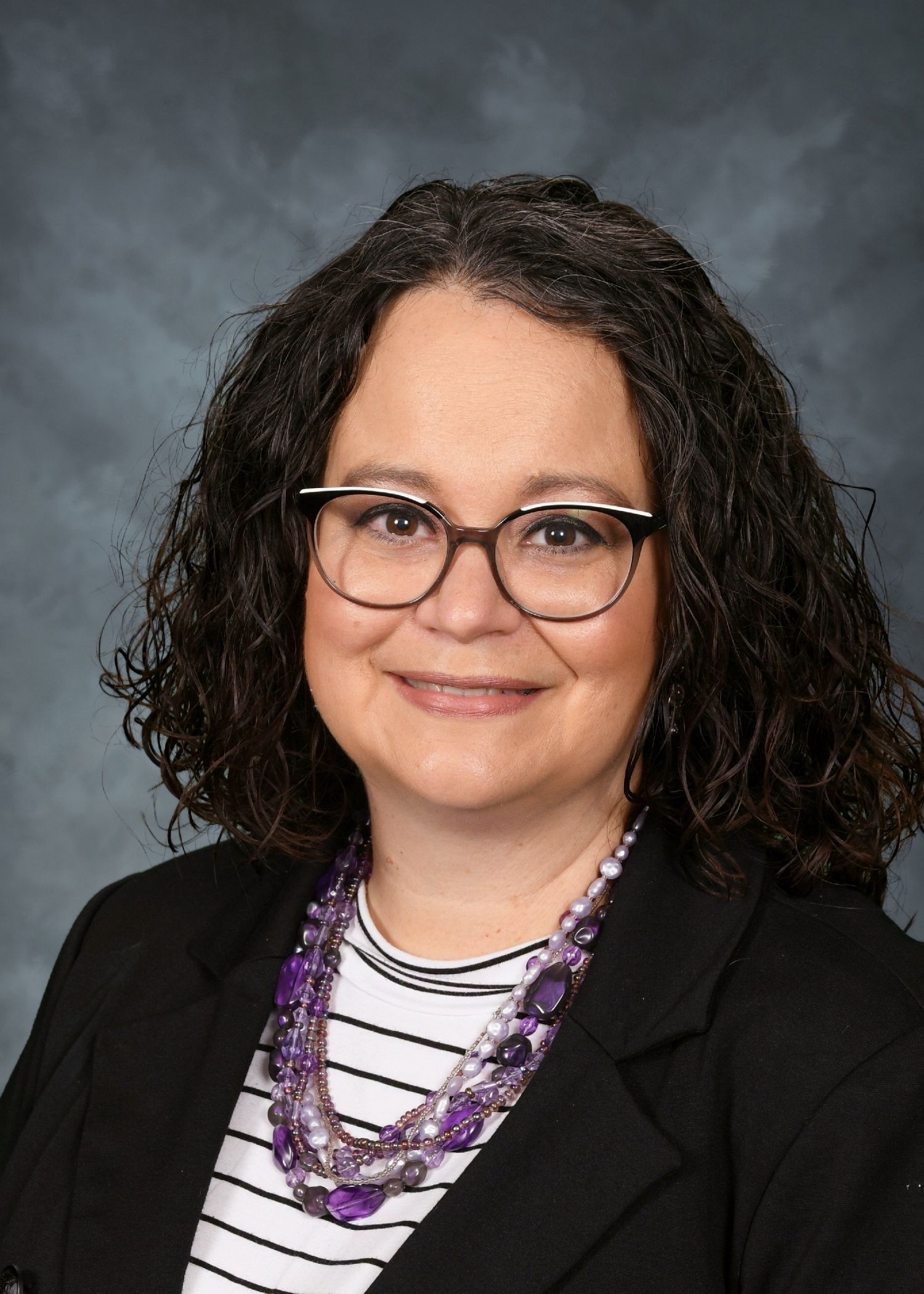 Woman wearing glasses, striped shirt, black jacket, and purple necklace, smiling.