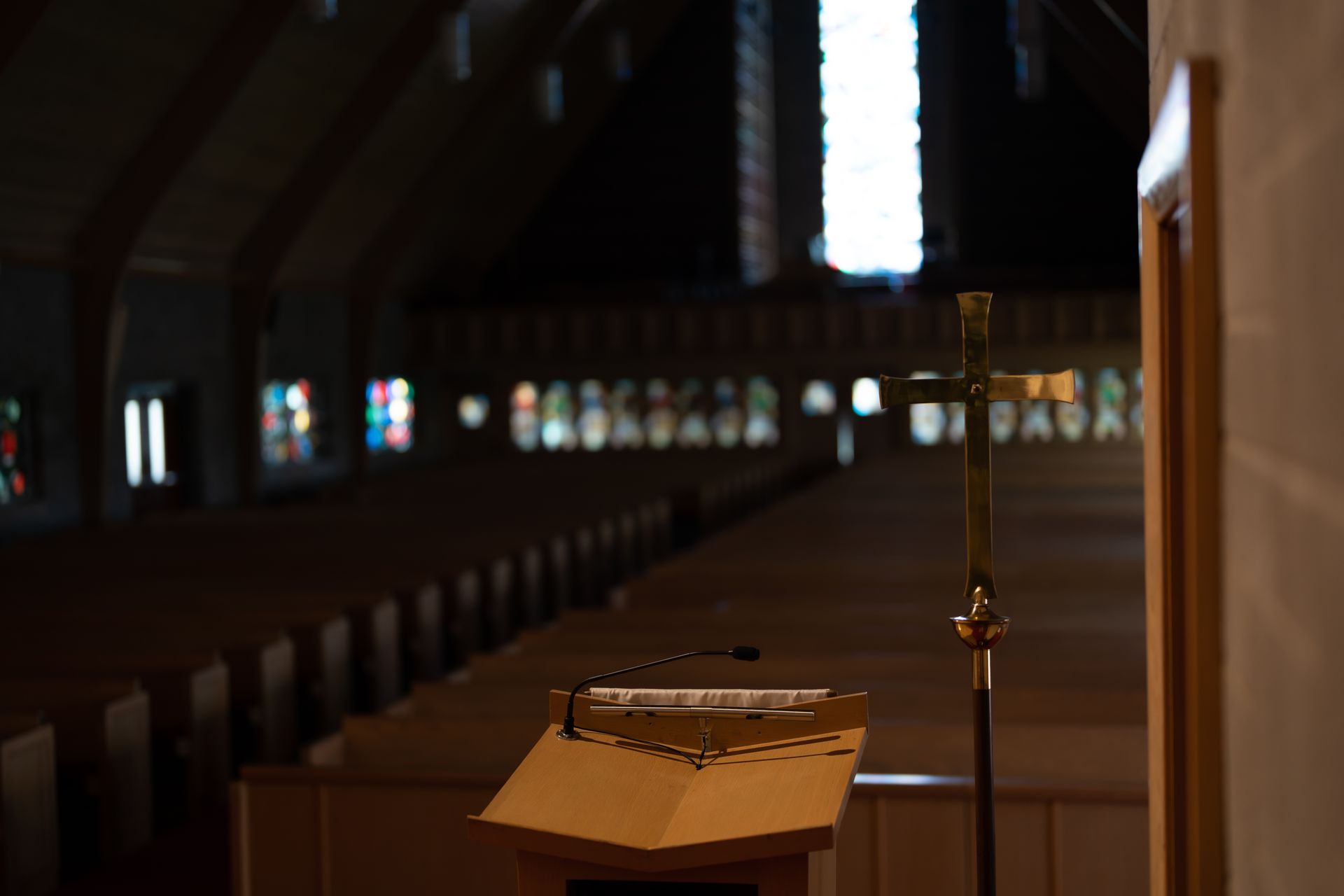Interior of a church with rows of pews, a podium, and a cross in the foreground. Sunlight streams through a tall window.