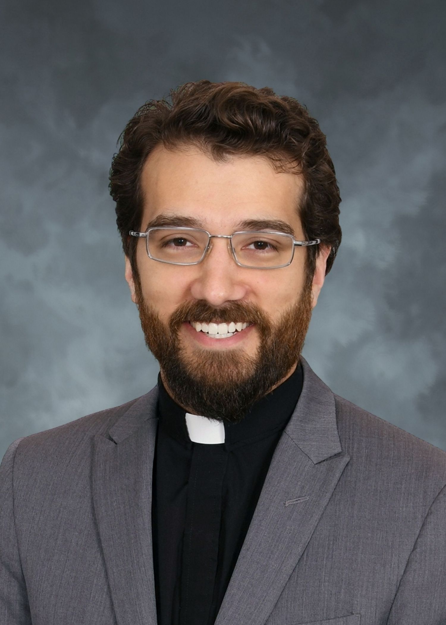 Man with glasses and beard, wearing clerical collar and gray blazer, smiles.