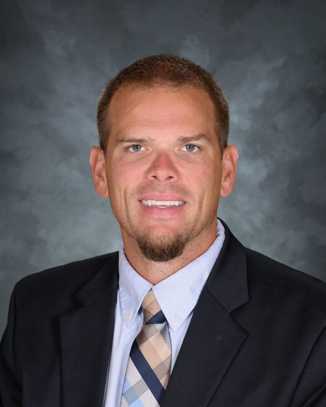 Man in navy blazer and patterned shirt smiles, headshot.