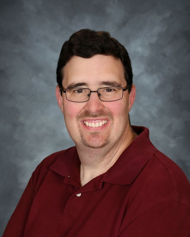 Man with glasses, wearing a light purple polo shirt, smiling, with a goatee, against a gray background.