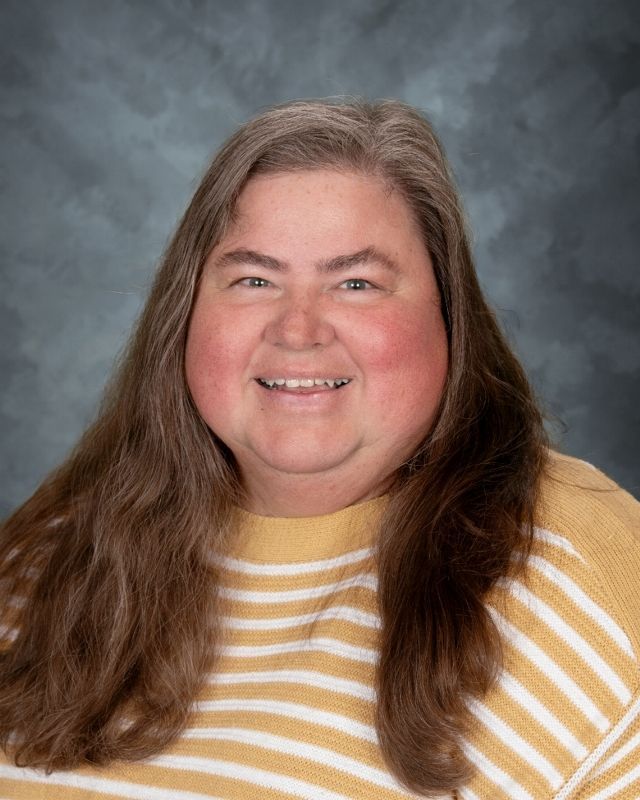 Woman with long brown hair smiles, wearing a navy floral print top. Background is a graduated blue and gray.