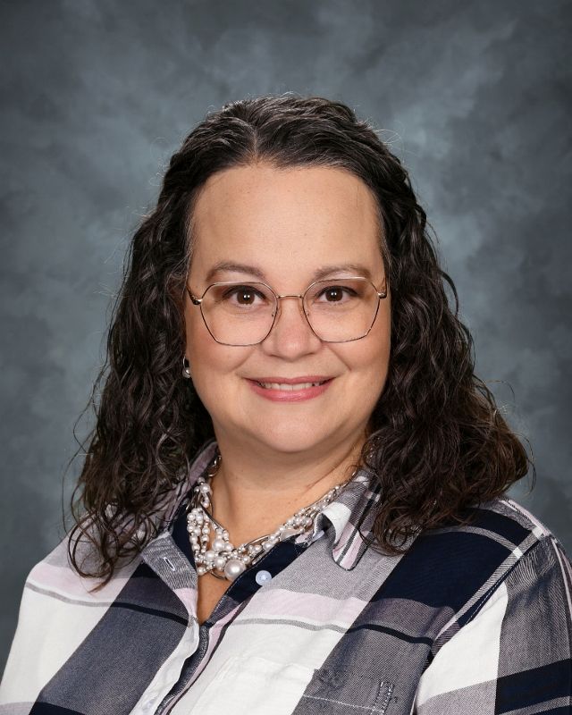 Woman with glasses, black jacket, and purple necklace smiles at the camera.