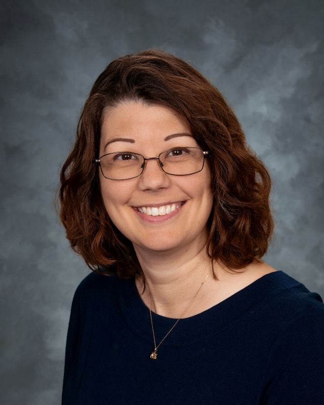Woman with glasses smiles; long curly brown hair, green shirt, dark gray background.