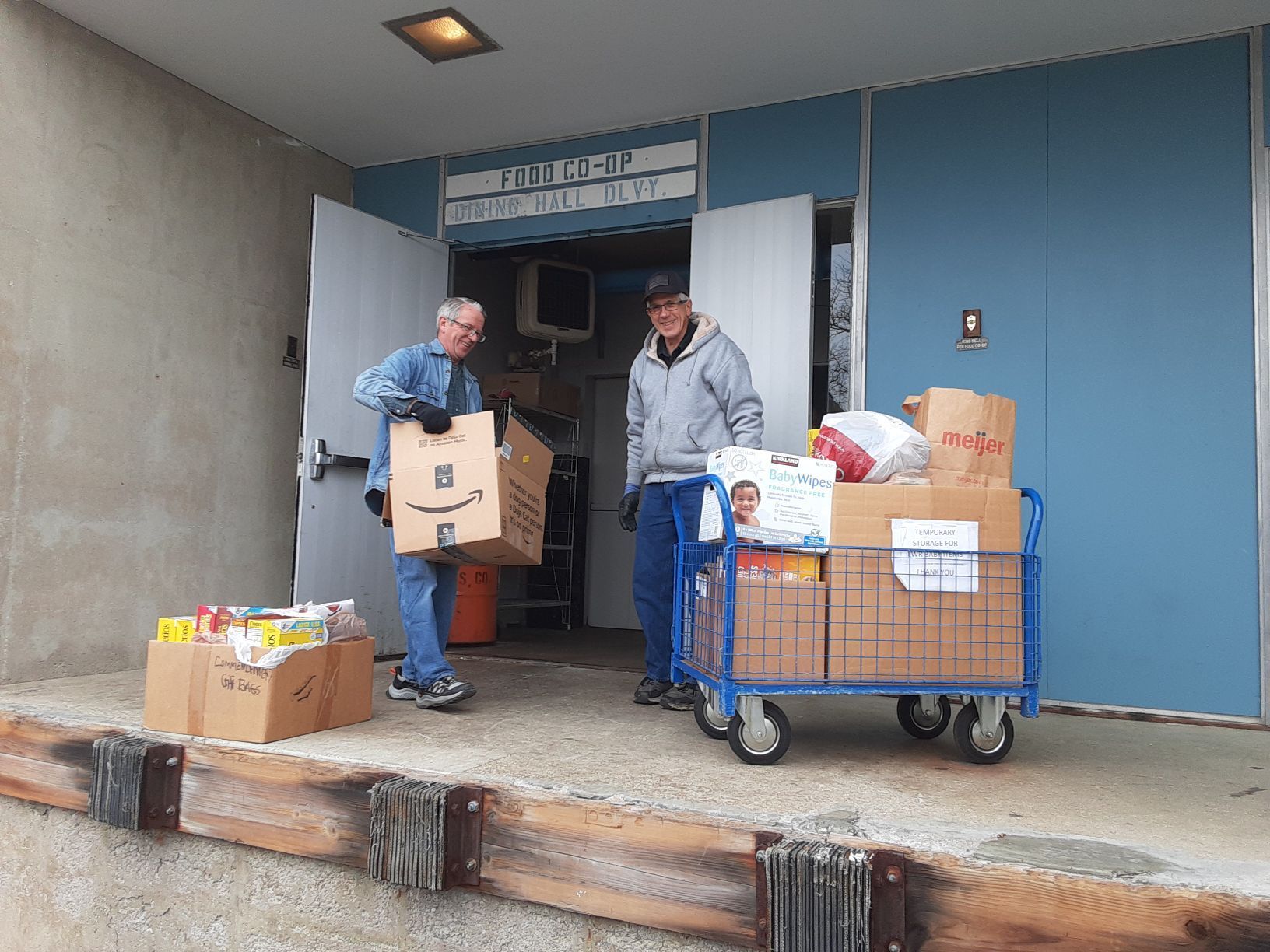 Two people unloading groceries from boxes and a cart at a building entrance.