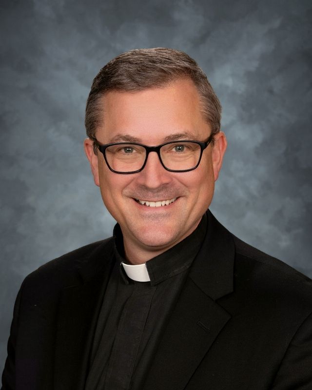 Man wearing glasses and clerical collar smiles.