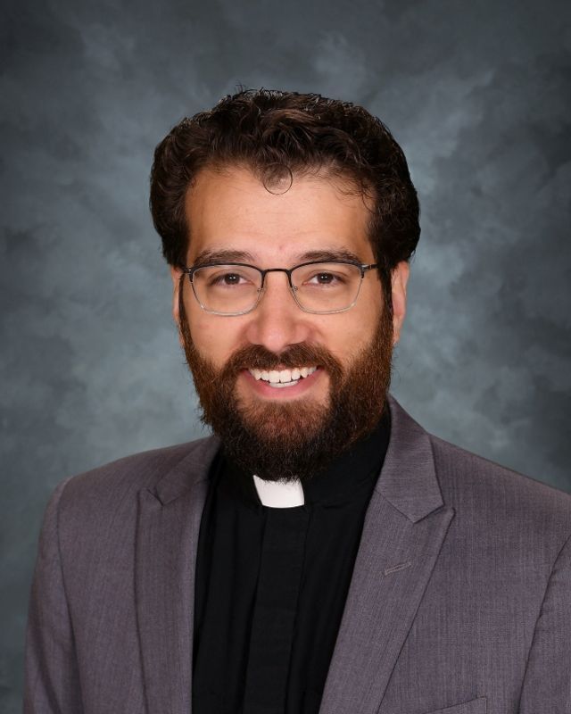 Man with glasses and beard, wearing a clerical collar and suit jacket, smiling, neutral background.