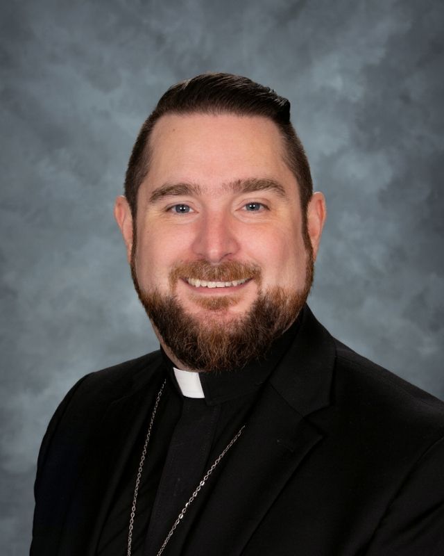 Clergyman with glasses, smiling, wearing a black suit and clerical collar.