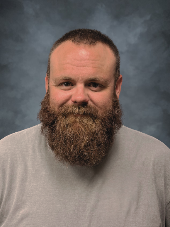 Man smiling in a blue polo shirt, against a blurred gray and blue background.