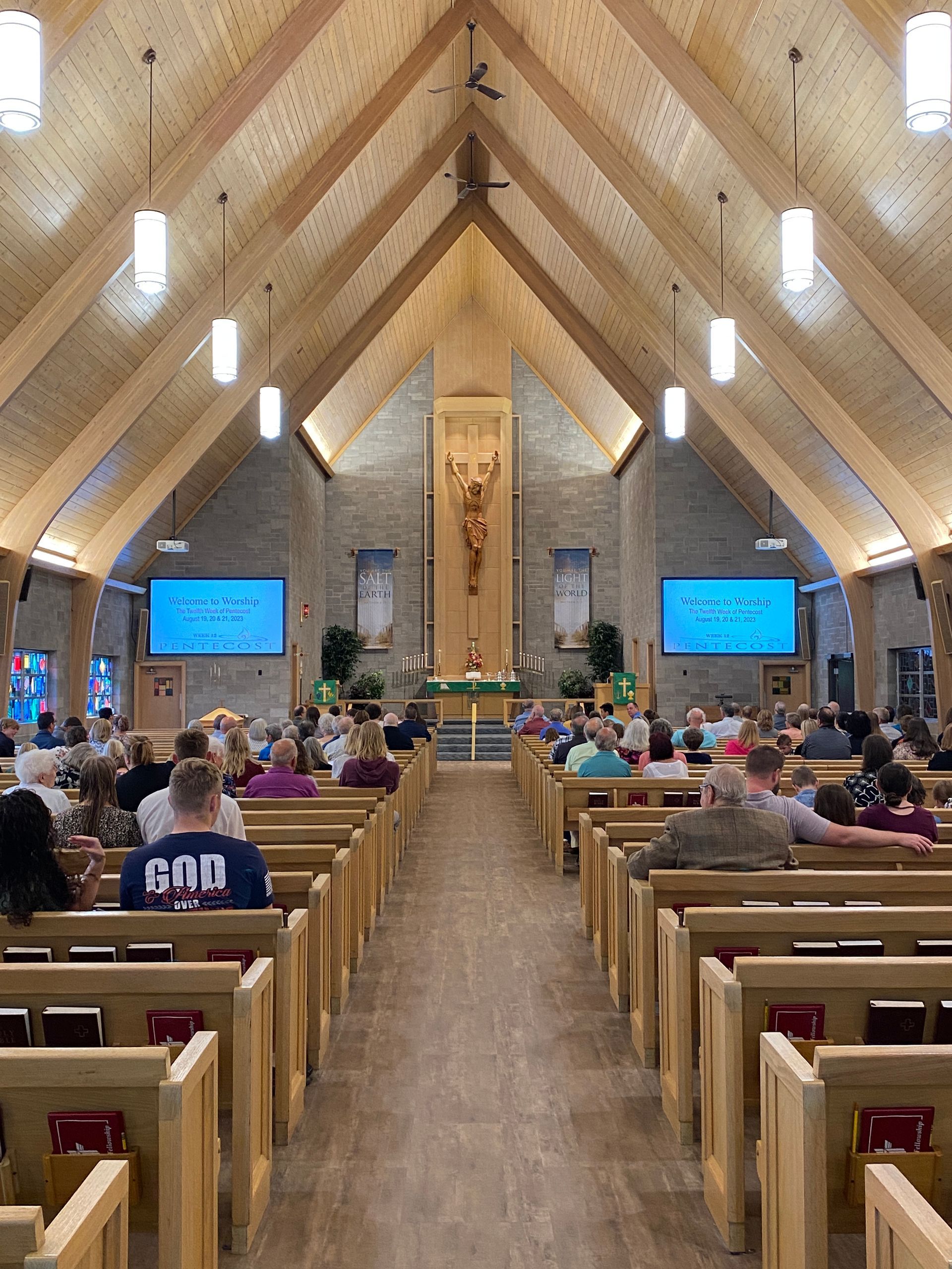 Interior of a church with people seated in pews, facing altar. Wooden beams on the ceiling, screens on the walls.