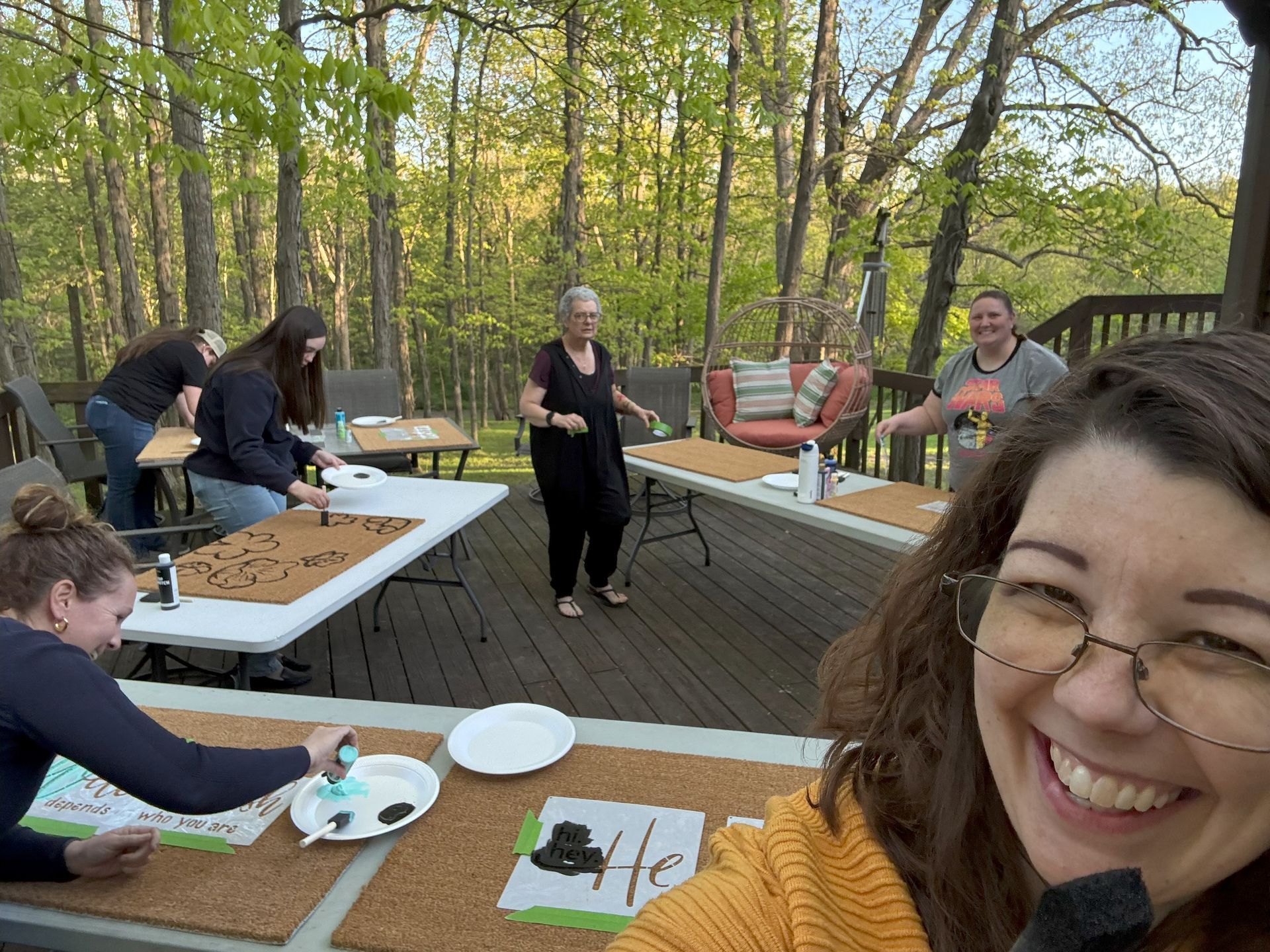 People painting doormats on an outdoor deck; smiling woman takes selfie. Tables, trees, and sunny day.
