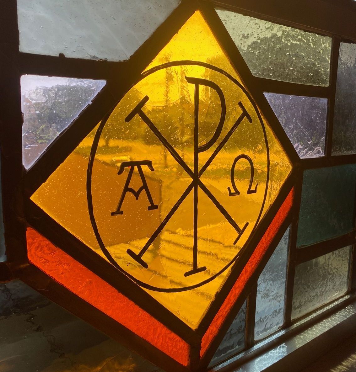 Stained glass window with a yellow diamond-shaped panel containing a Chi Rho symbol and the Greek letters Alpha and Omega.