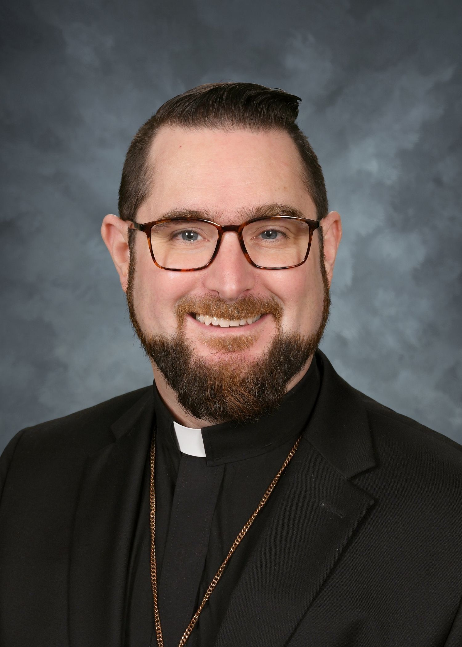 Clerical man with glasses and beard, wearing a black suit and collar, smiling.