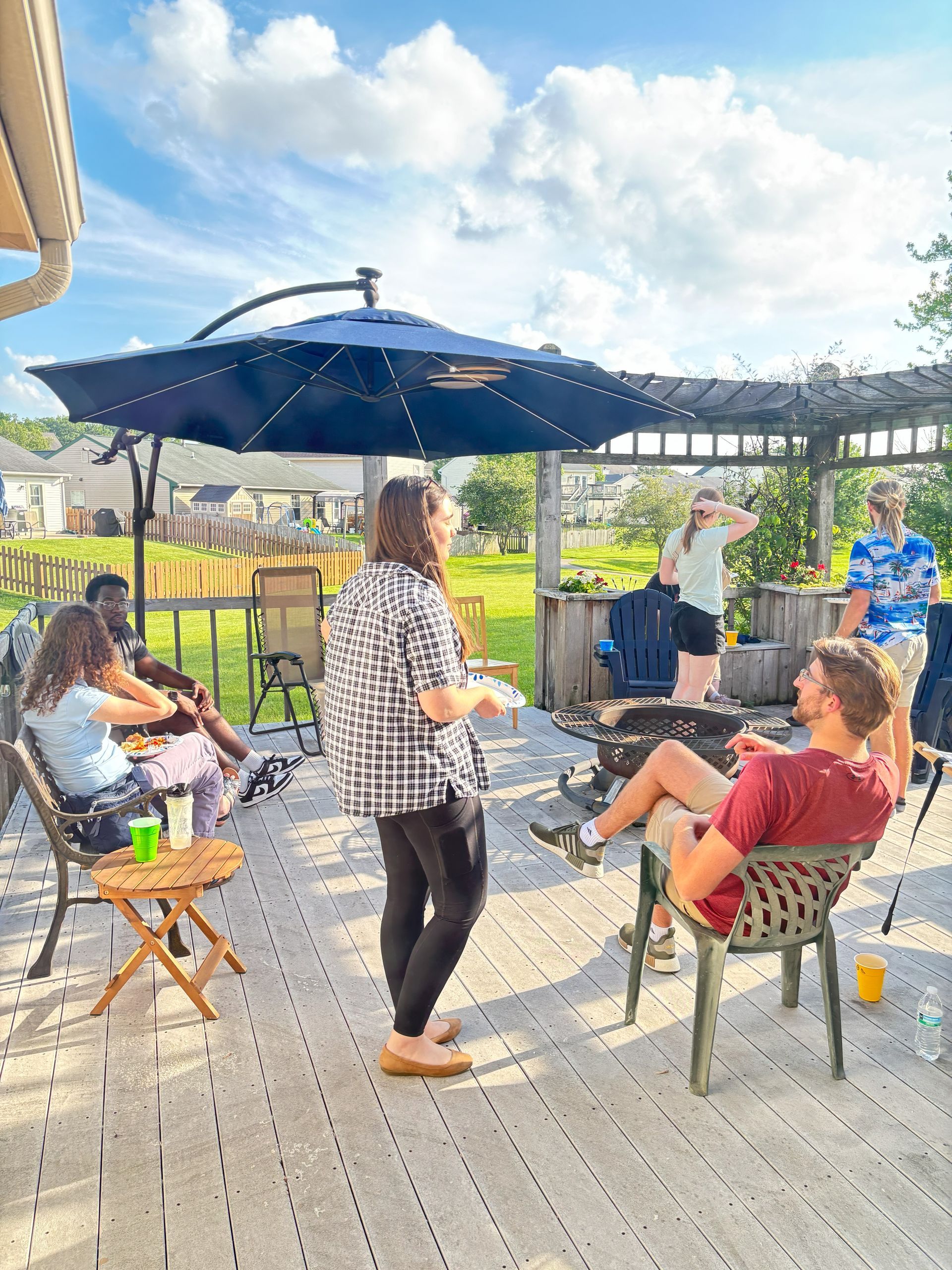 Group of people socializing on a sunny deck under an umbrella. People are seated and standing, talking.