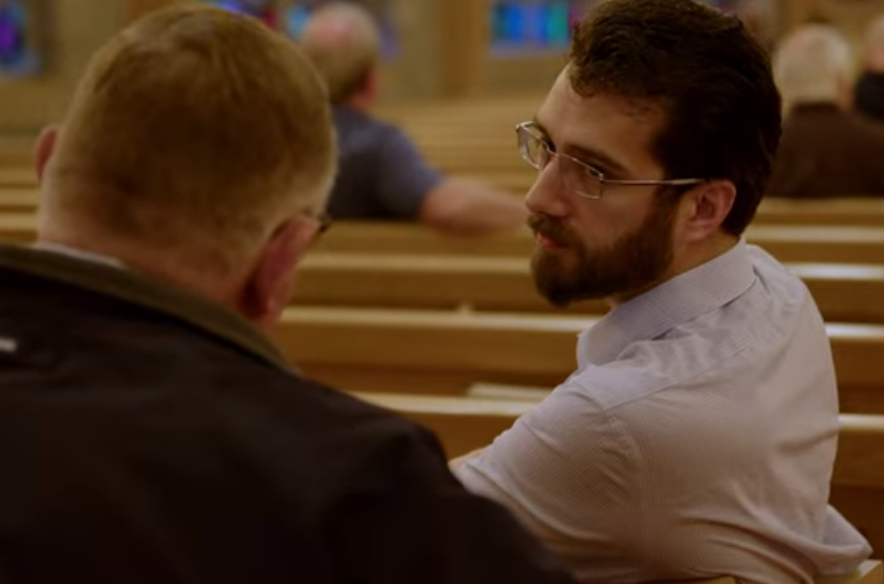 Two men in a church, one with glasses and a beard, talking while seated on a wooden pew.