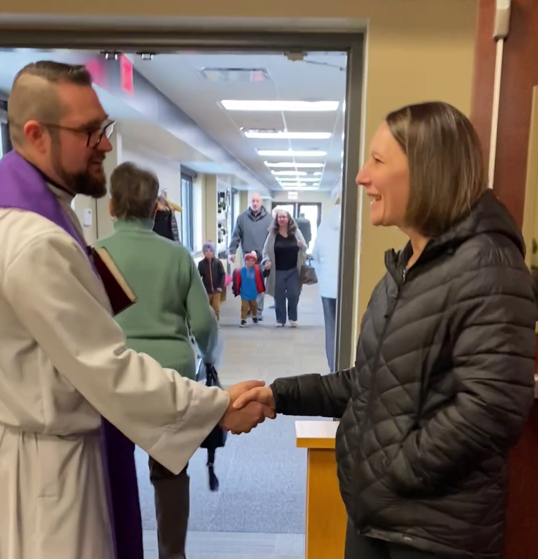 A person in clerical collar shakes hands with a woman; hallway with people in background.