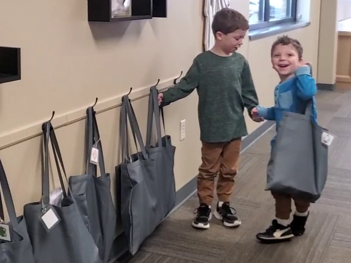 Two boys holding hands, smiling, next to tote bags hanging on wall hooks.