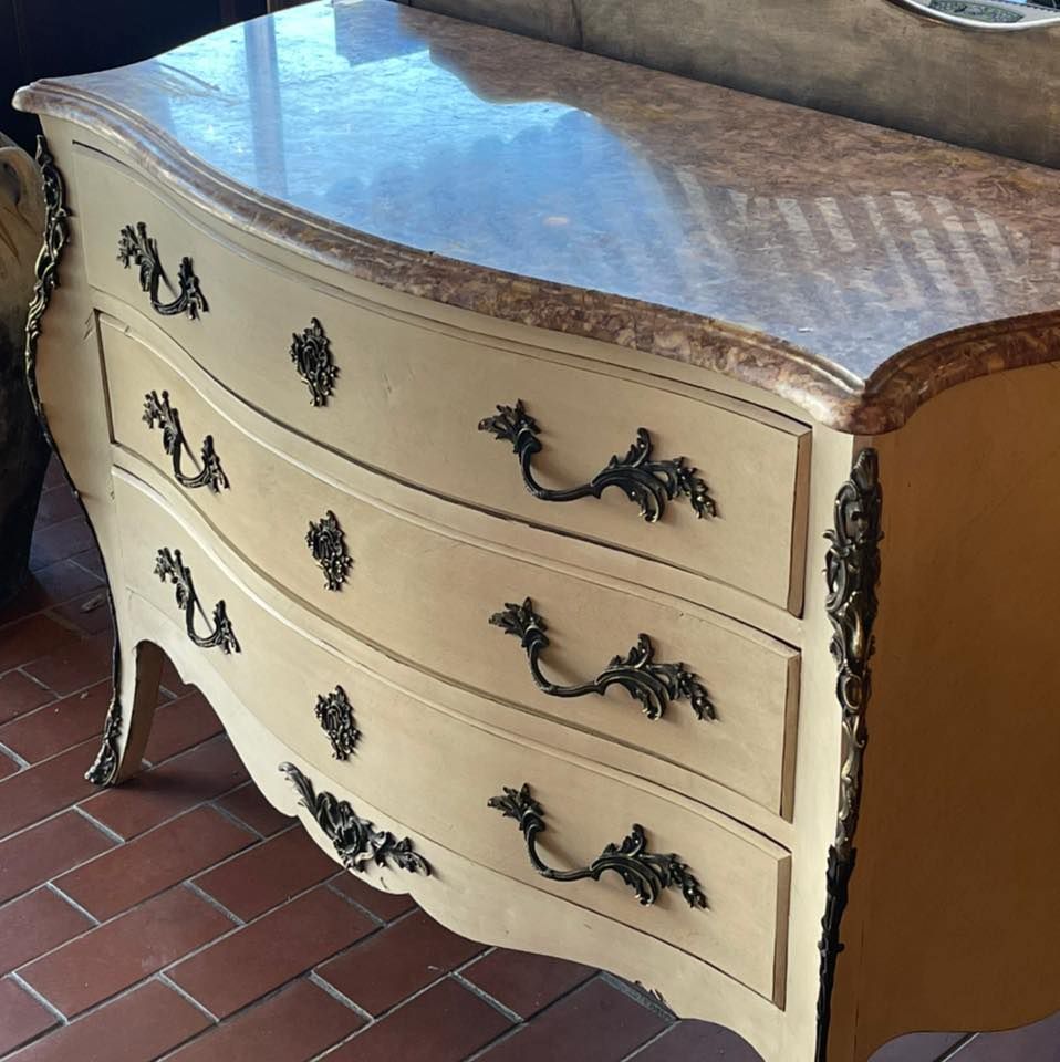 Cream-colored antique chest of drawers with curved front, bronze hardware, and marble top.