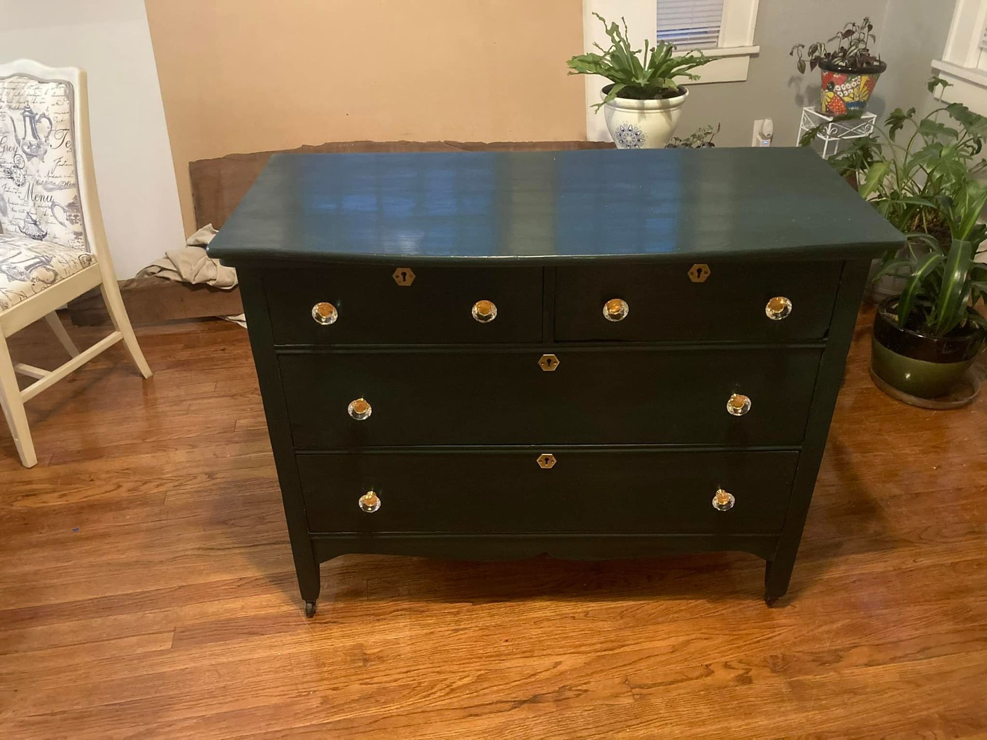 Dark green dresser with gold knobs on a wooden floor, next to a chair and plants.