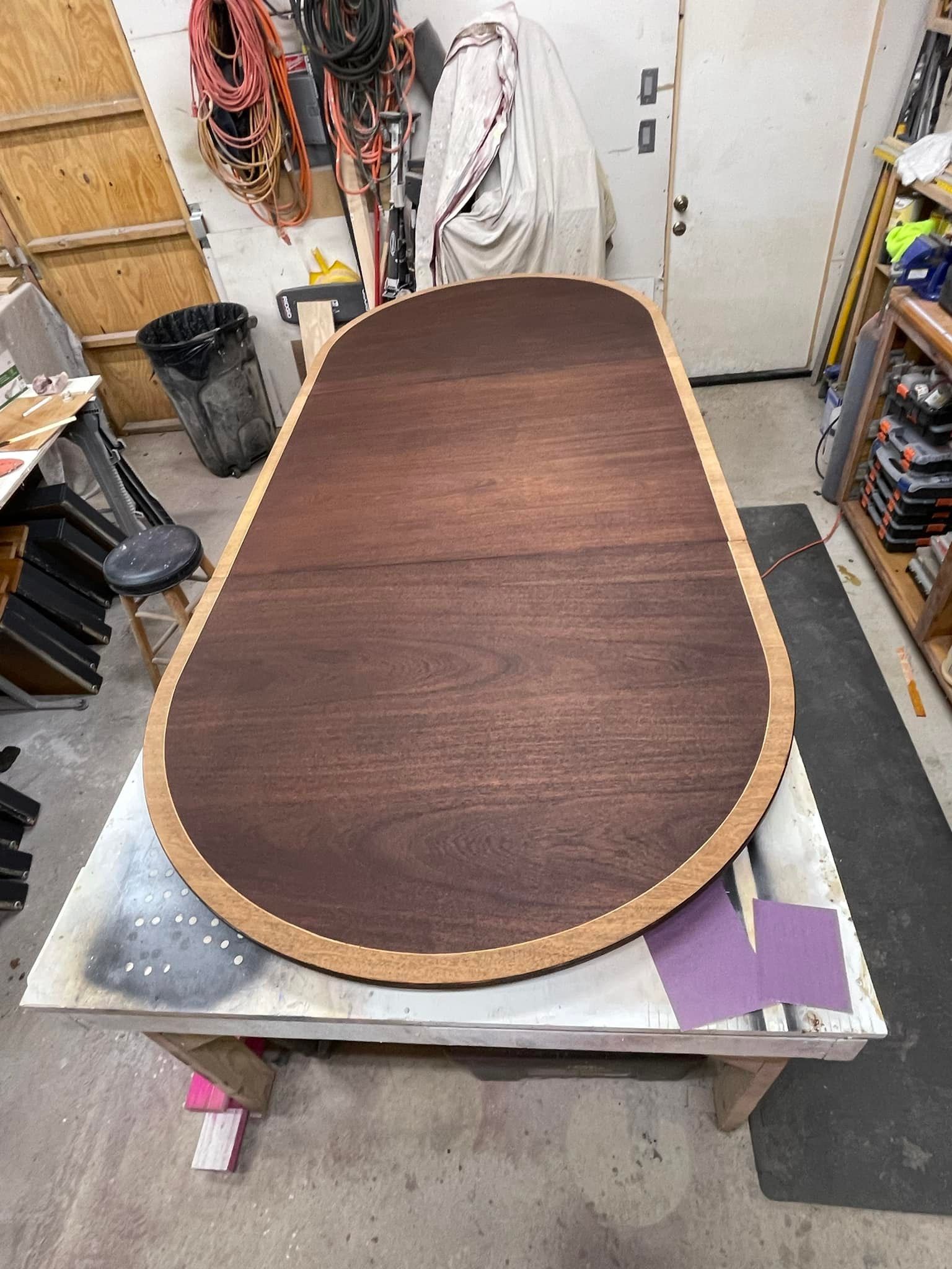 A wood oval table with a dark brown center and a light brown border in a workshop.