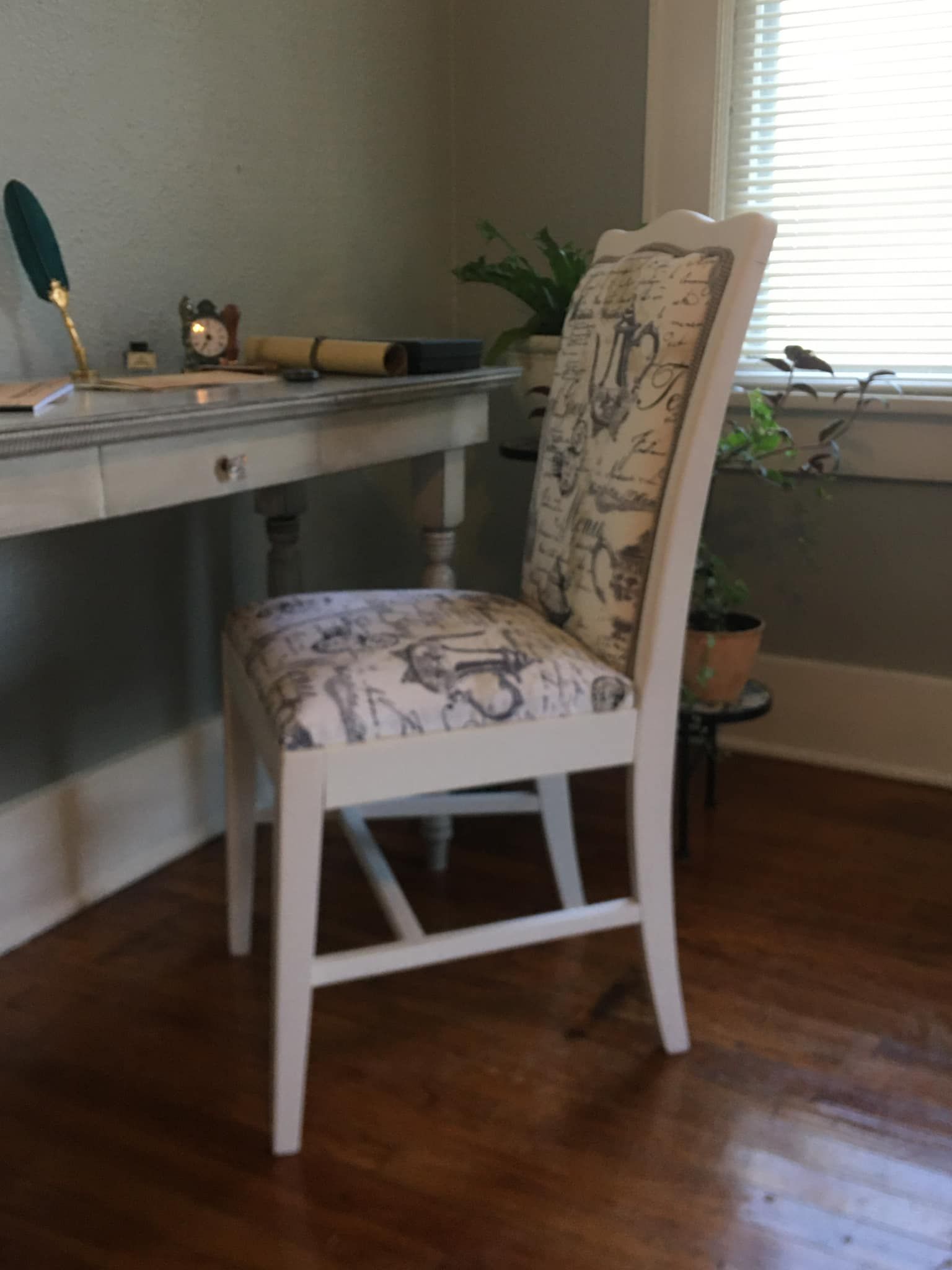 White painted chair with fabric seat at a writing desk, next to a window.