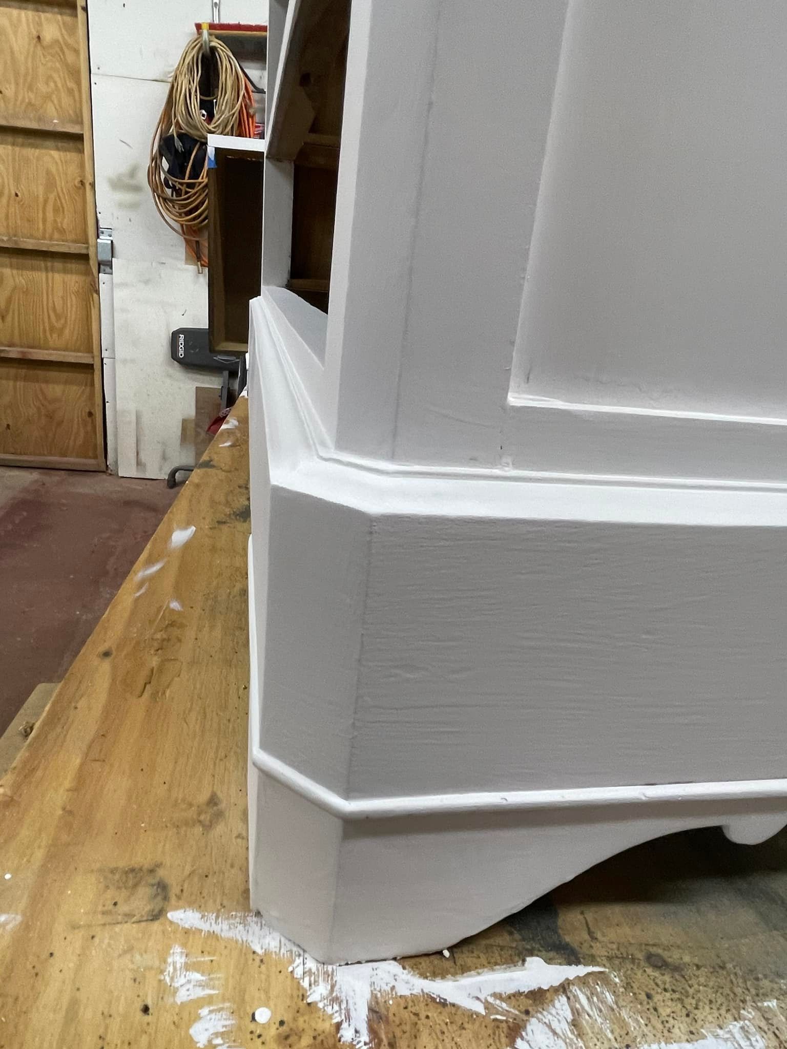 Close-up of a white painted cabinet corner with trim details, sitting on a wooden surface.