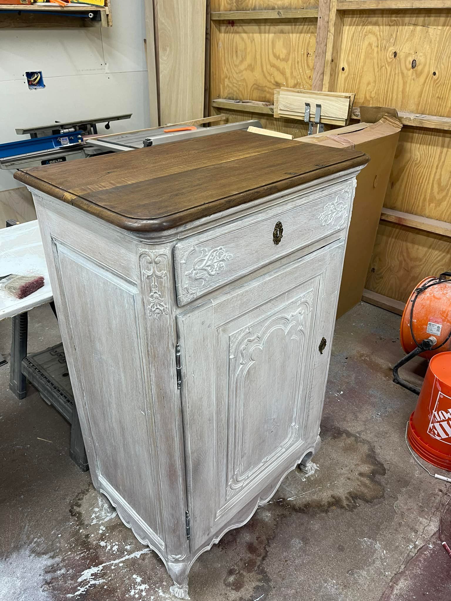 Antique cabinet with detailed carving, painted white with stained wood top, in a workshop.