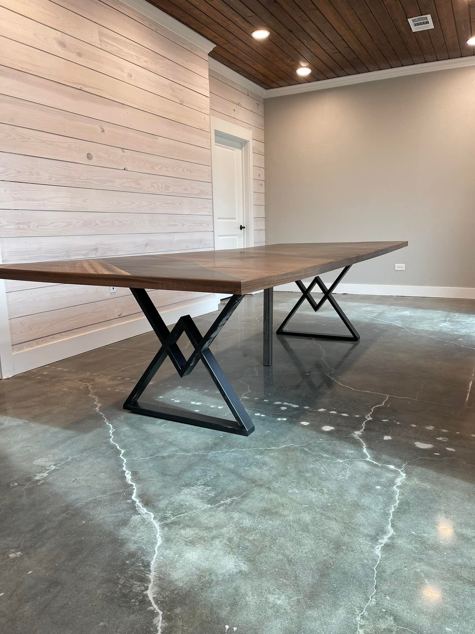 Large wooden table with black metal legs in a room with a concrete floor, light gray walls, and wood-paneled ceiling.