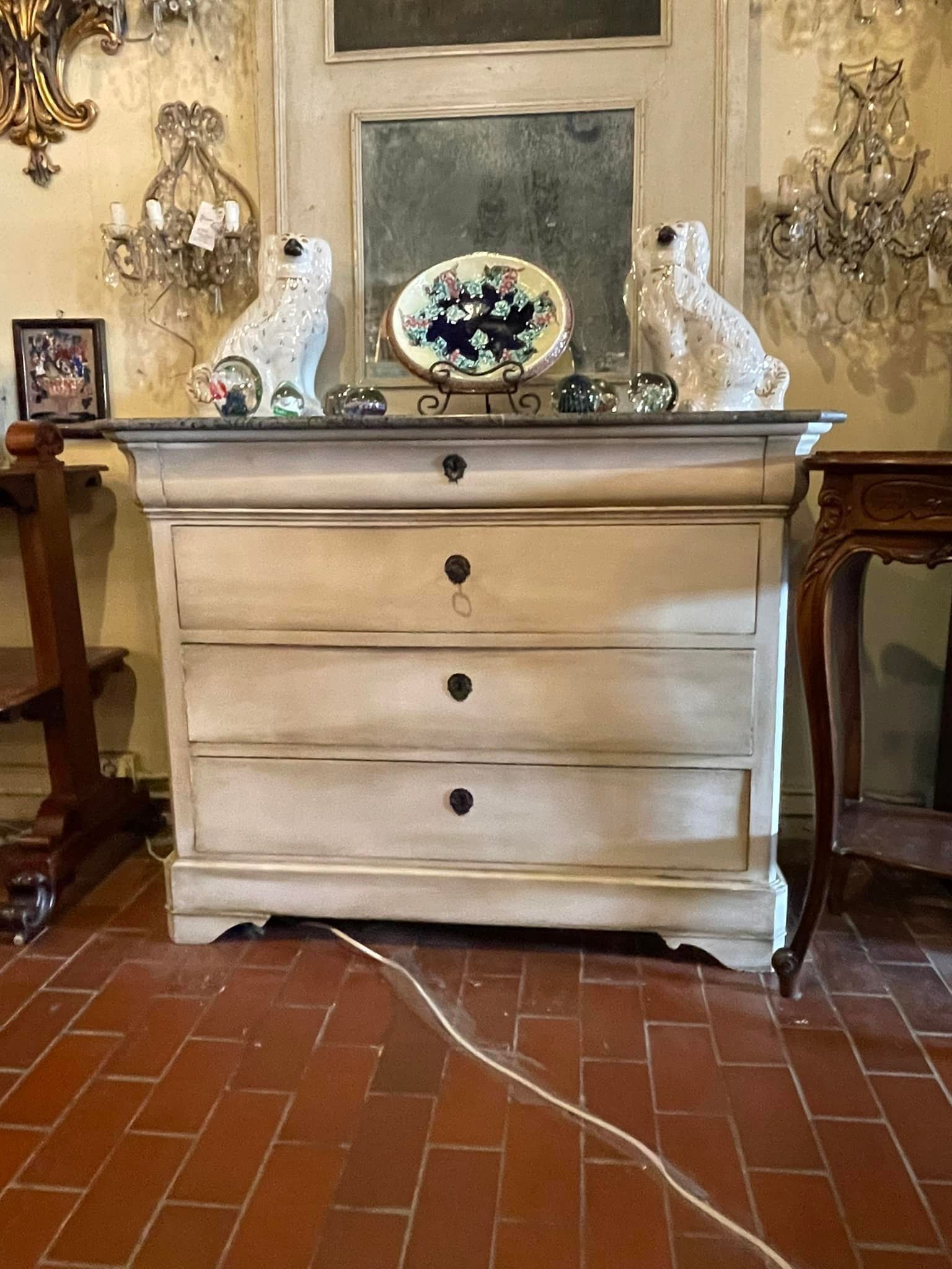 Cream-colored chest of drawers with three drawers, topped with dog statues and a decorative plate, set on a red-tiled floor.