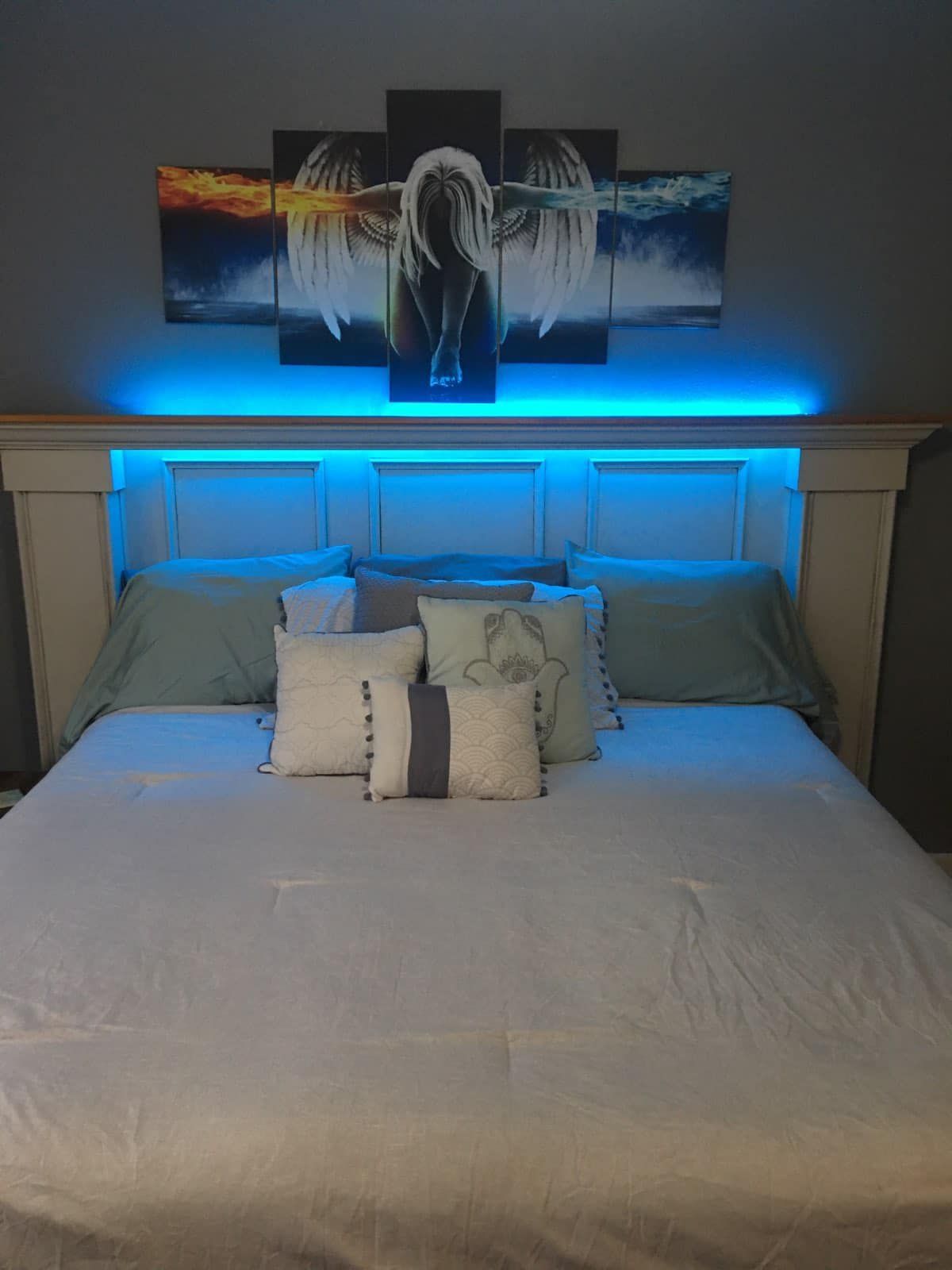 Bed with a white headboard, blue LED lighting, and artwork above it; pillows and a light-colored blanket.