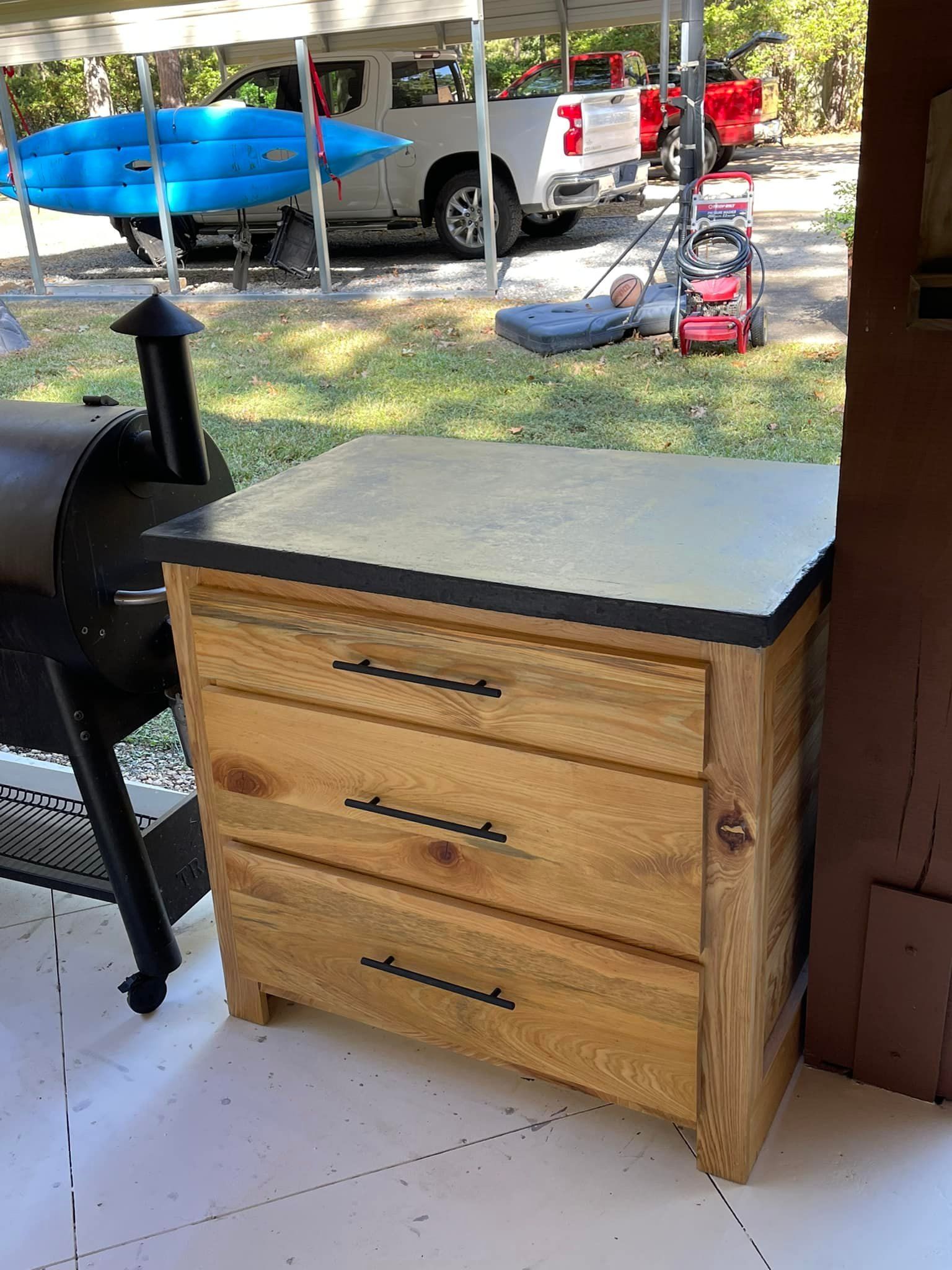 Wooden cabinet with three drawers and a black countertop, next to a smoker.
