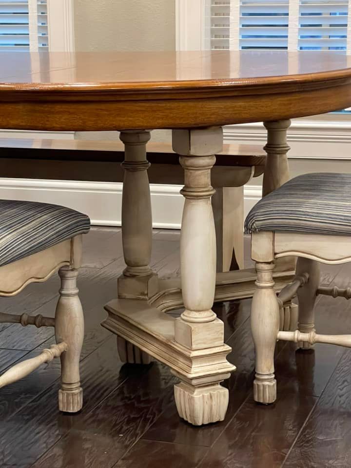 Wooden dining table with painted legs and two chairs. Tabletop is brown, legs are off-white, chairs have blue-striped cushions.