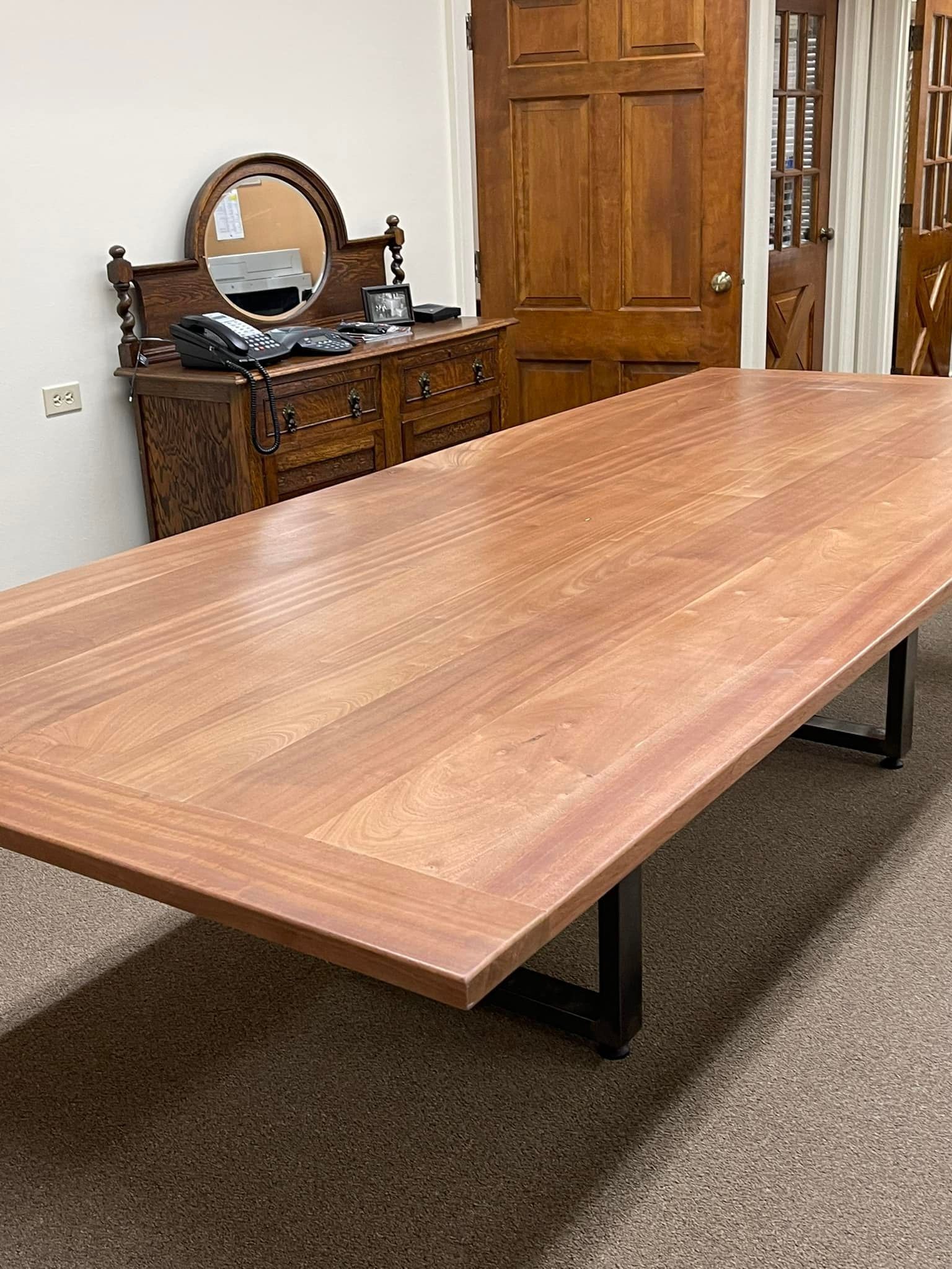 A long wooden table with black metal legs, against a wall. A dresser and door are in the background.