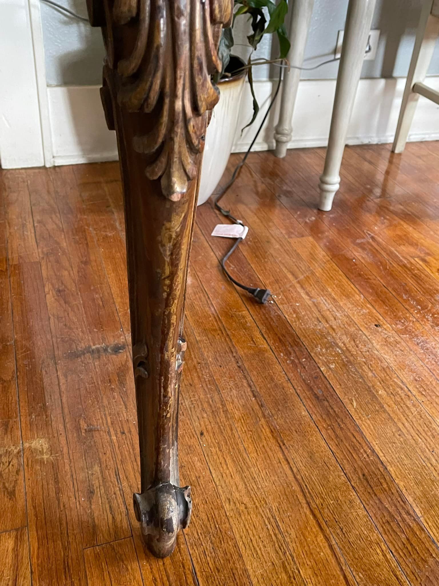 Ornate wooden table leg, with carvings, on hardwood floor. A power cord is visible nearby.