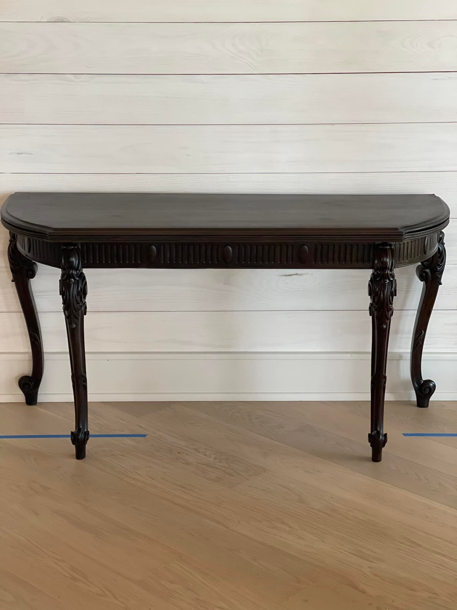 Dark brown, half-moon shaped console table with carved details and curved legs against white shiplap and wood floor.