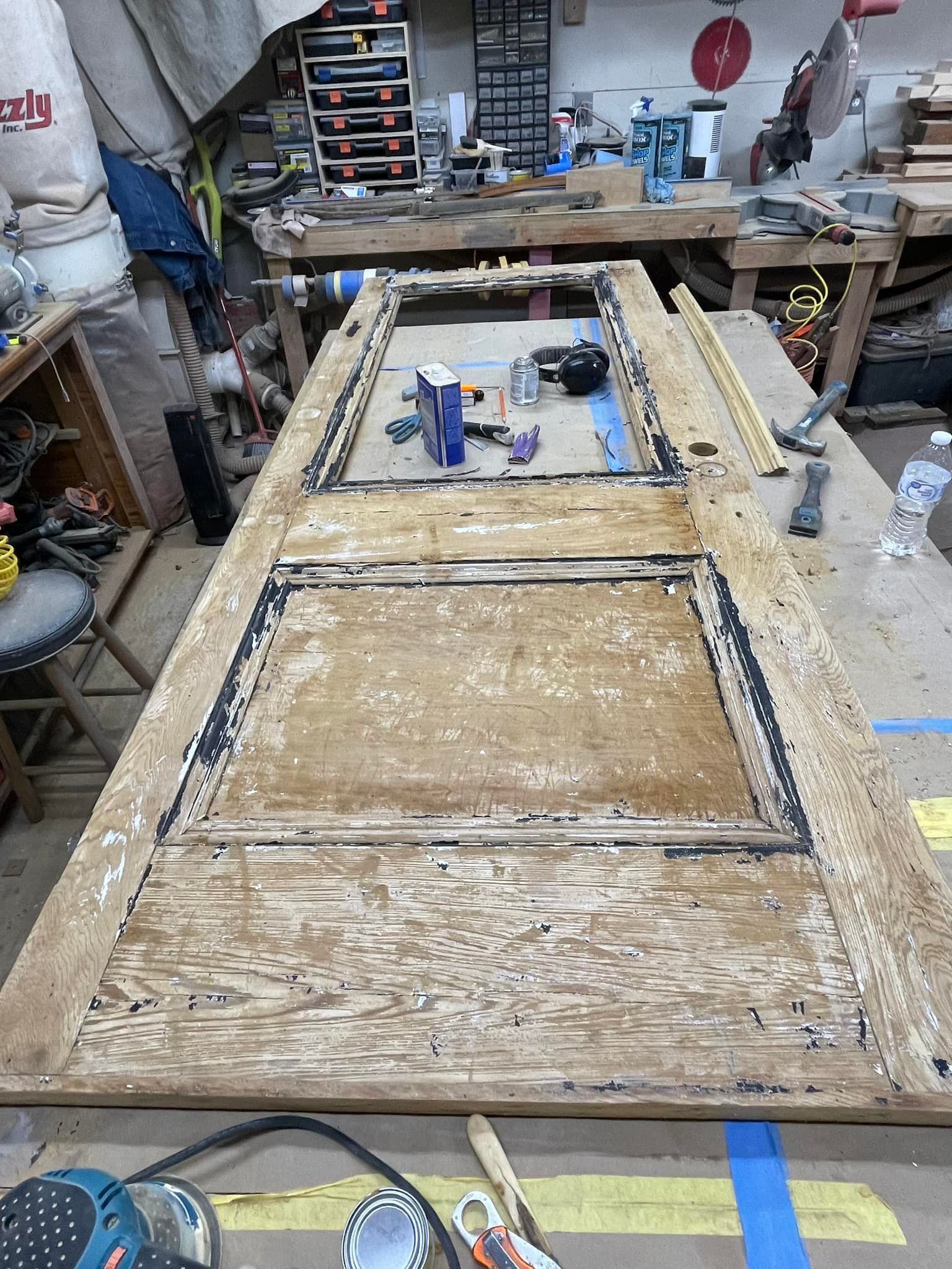 Wooden door laid flat on a workbench, undergoing repair. Surrounding workshop with tools and materials.