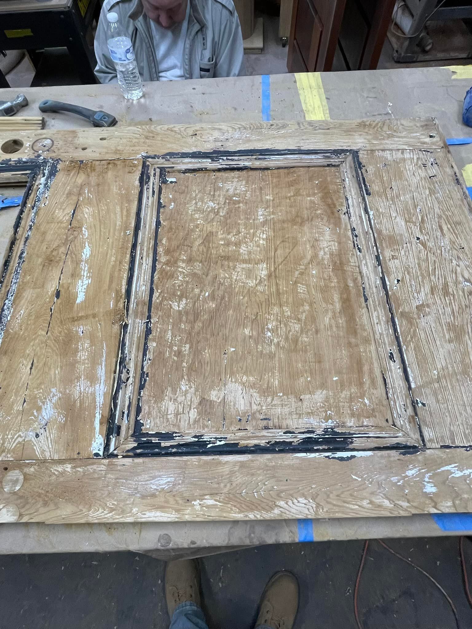 A wood door being worked on in a workshop; a person is visible. Door has peeling paint.