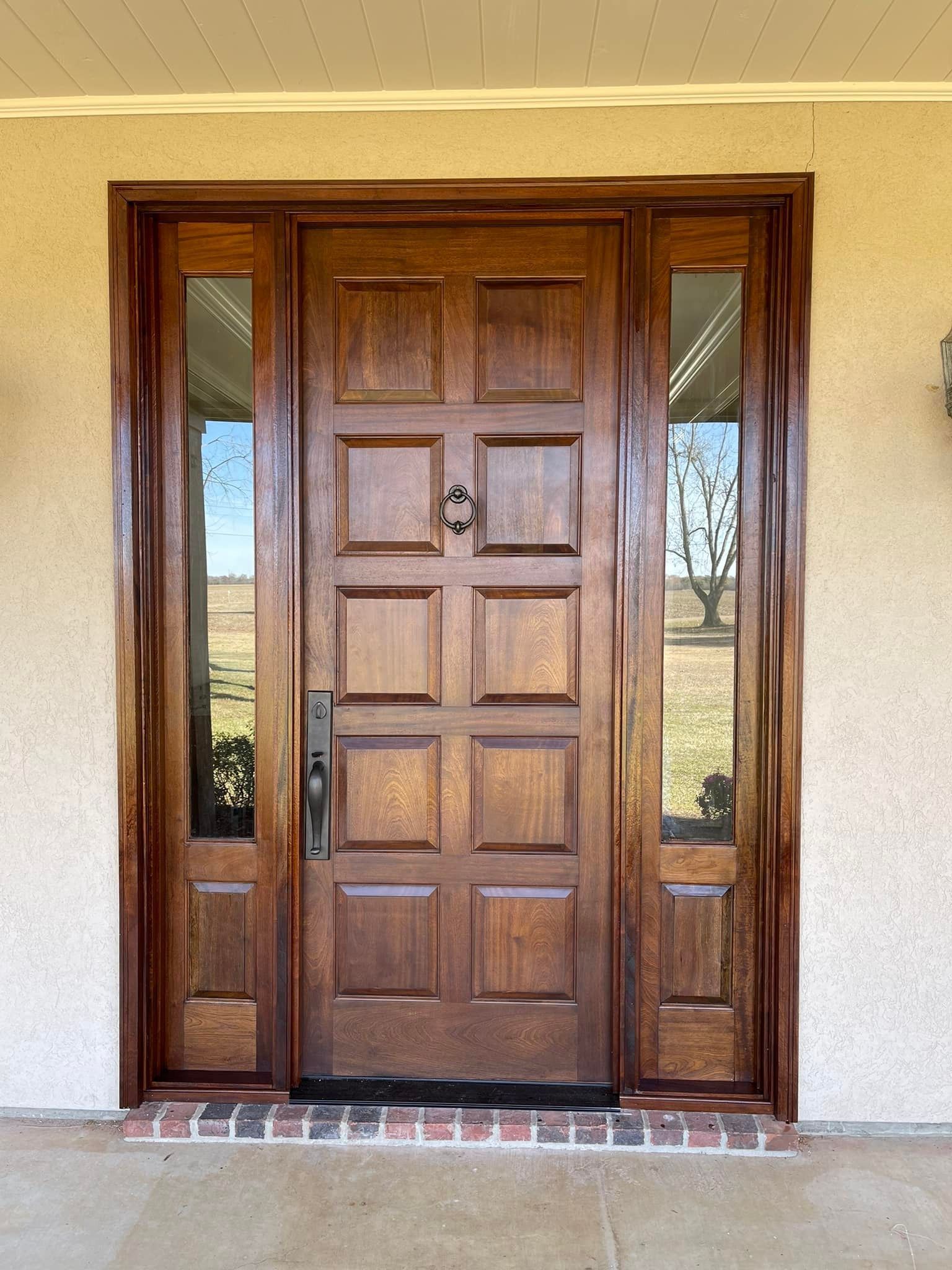 Wooden front door with sidelights, brown color, on a tan building with a brick threshold.