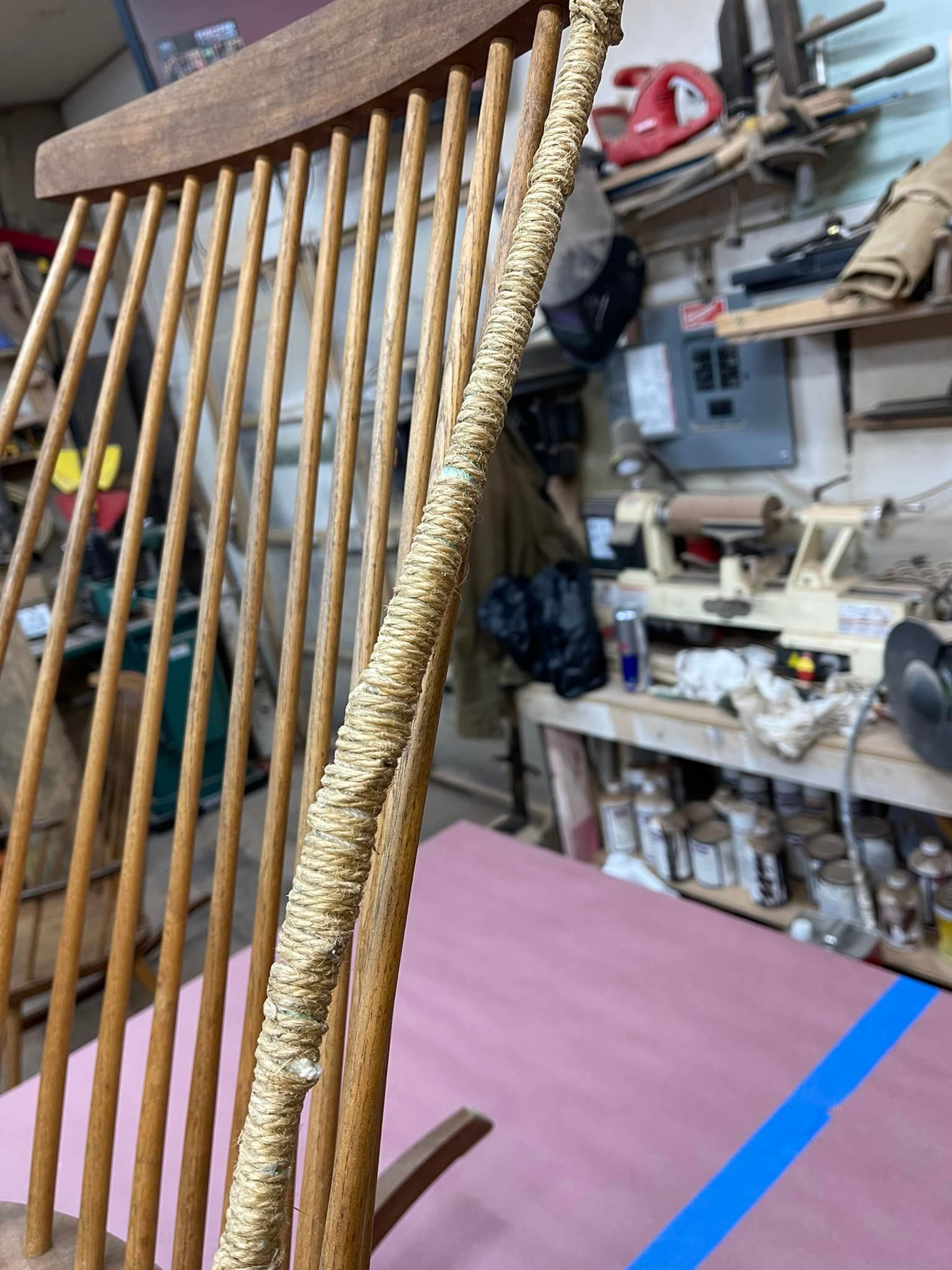 Close-up of a wooden chair back with a textured, wrapped piece along one of the vertical supports in a workshop setting.