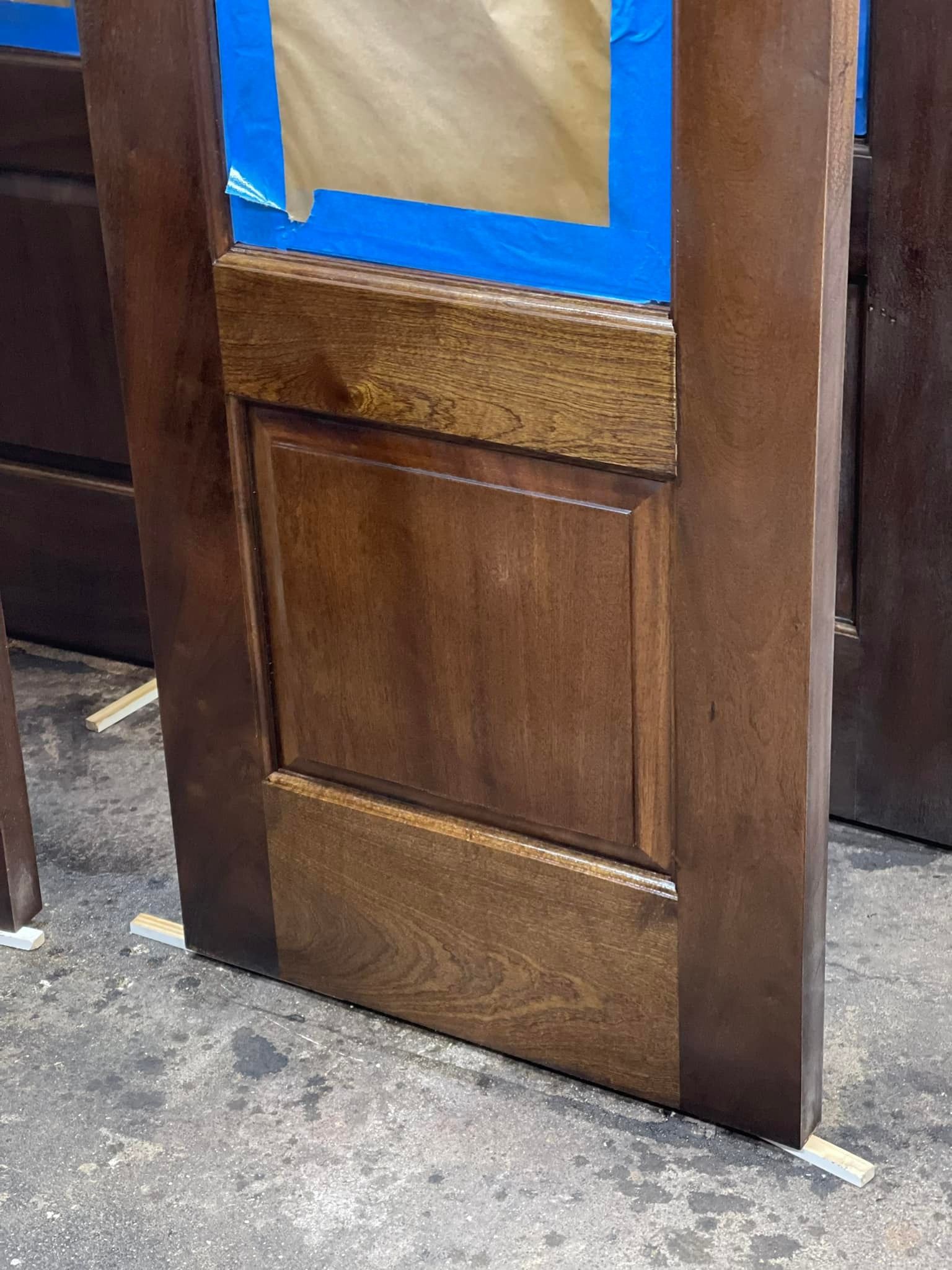 Wood door with stained panels and blue painter's tape; standing upright in a workshop setting.