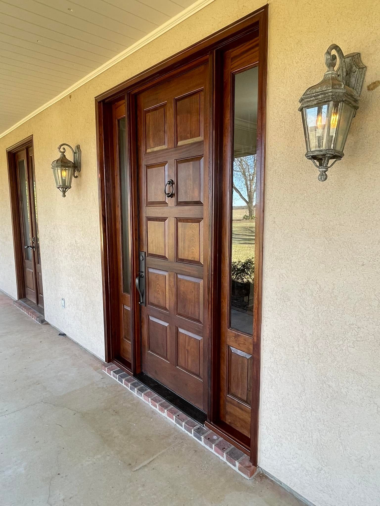 Wooden front door with sidelights, flanked by ornate wall sconces on a stucco exterior.
