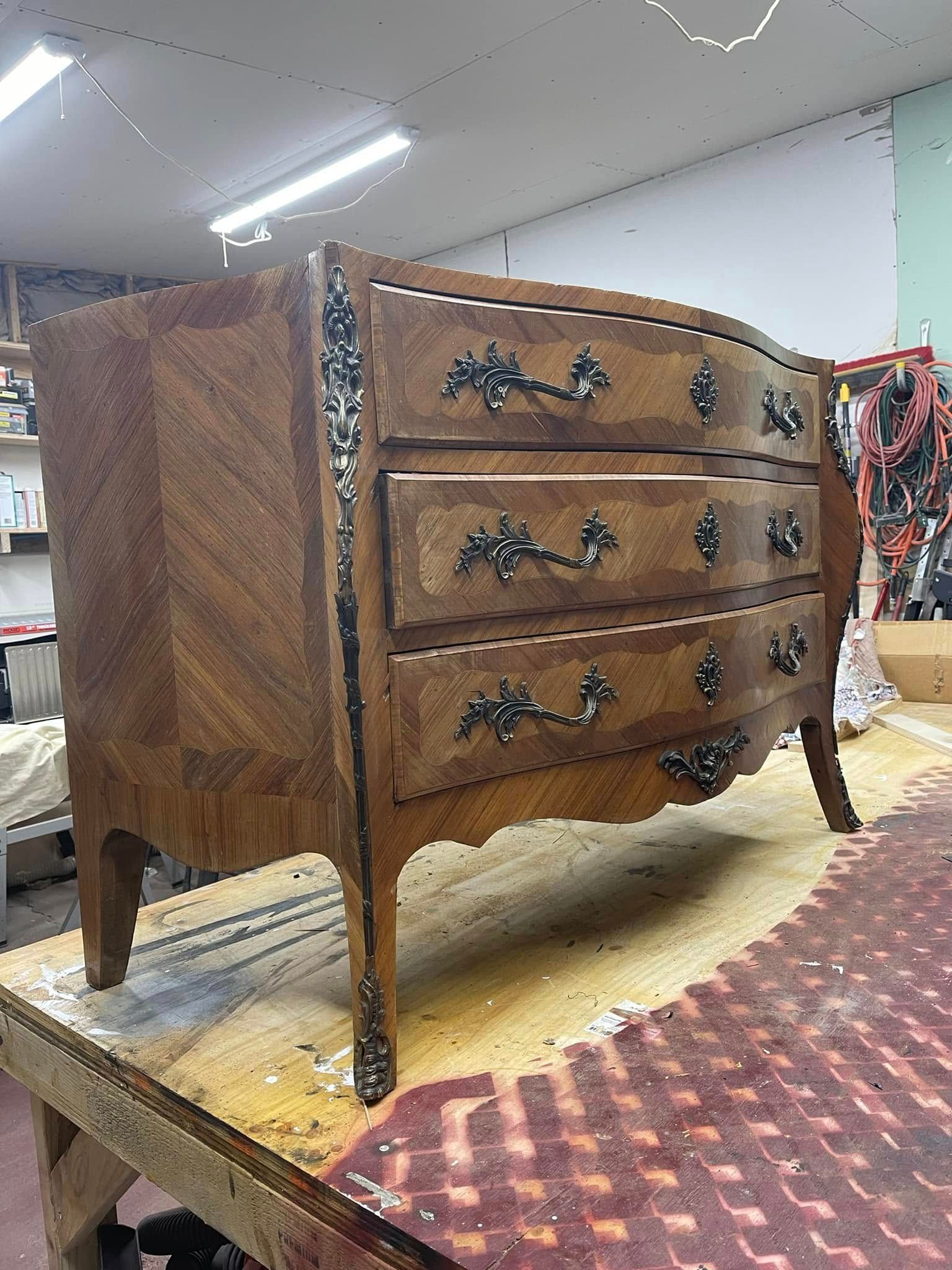 A wooden, three-drawer dresser with ornate hardware and legs, sits on a workbench.