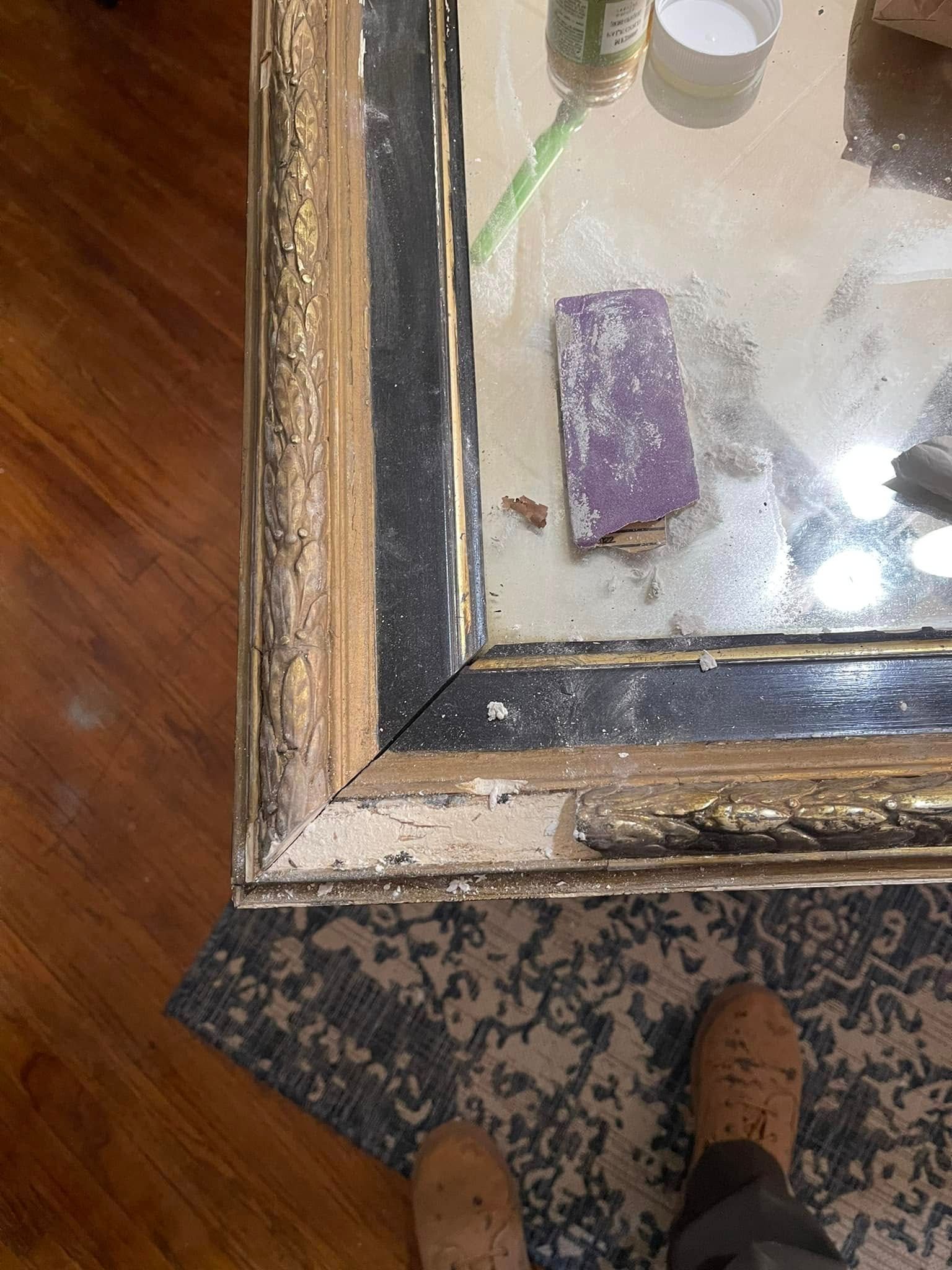 Close-up of a damaged gold and black framed mirror with a sanding block on the reflective surface.
