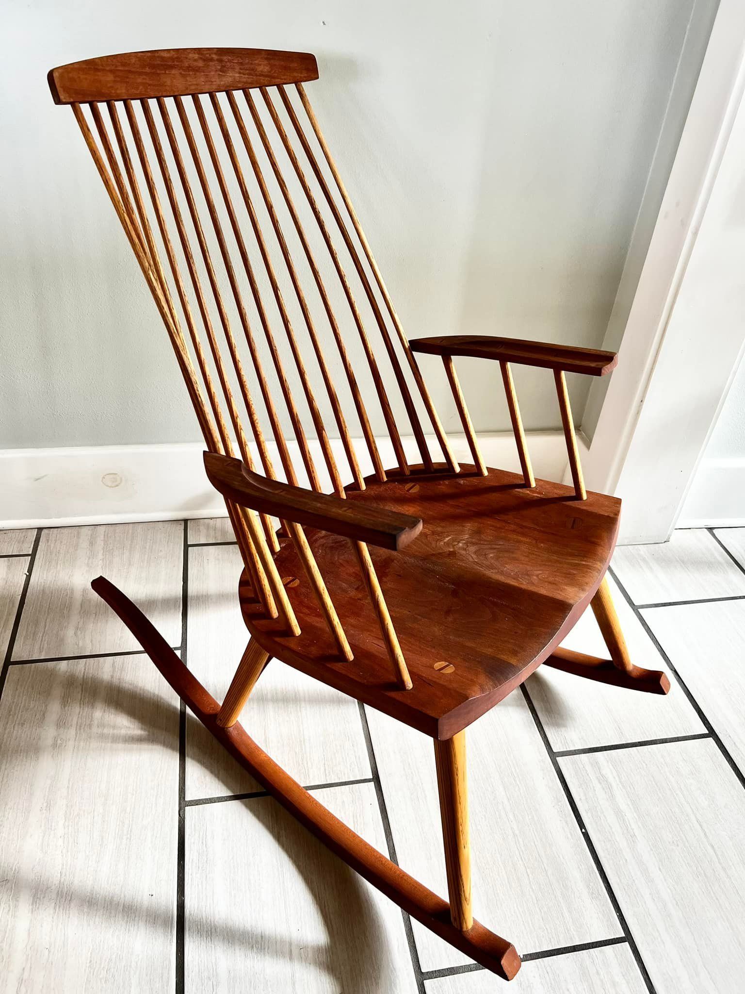 Wooden rocking chair with a tall, slatted back and armrests, on a tiled floor.