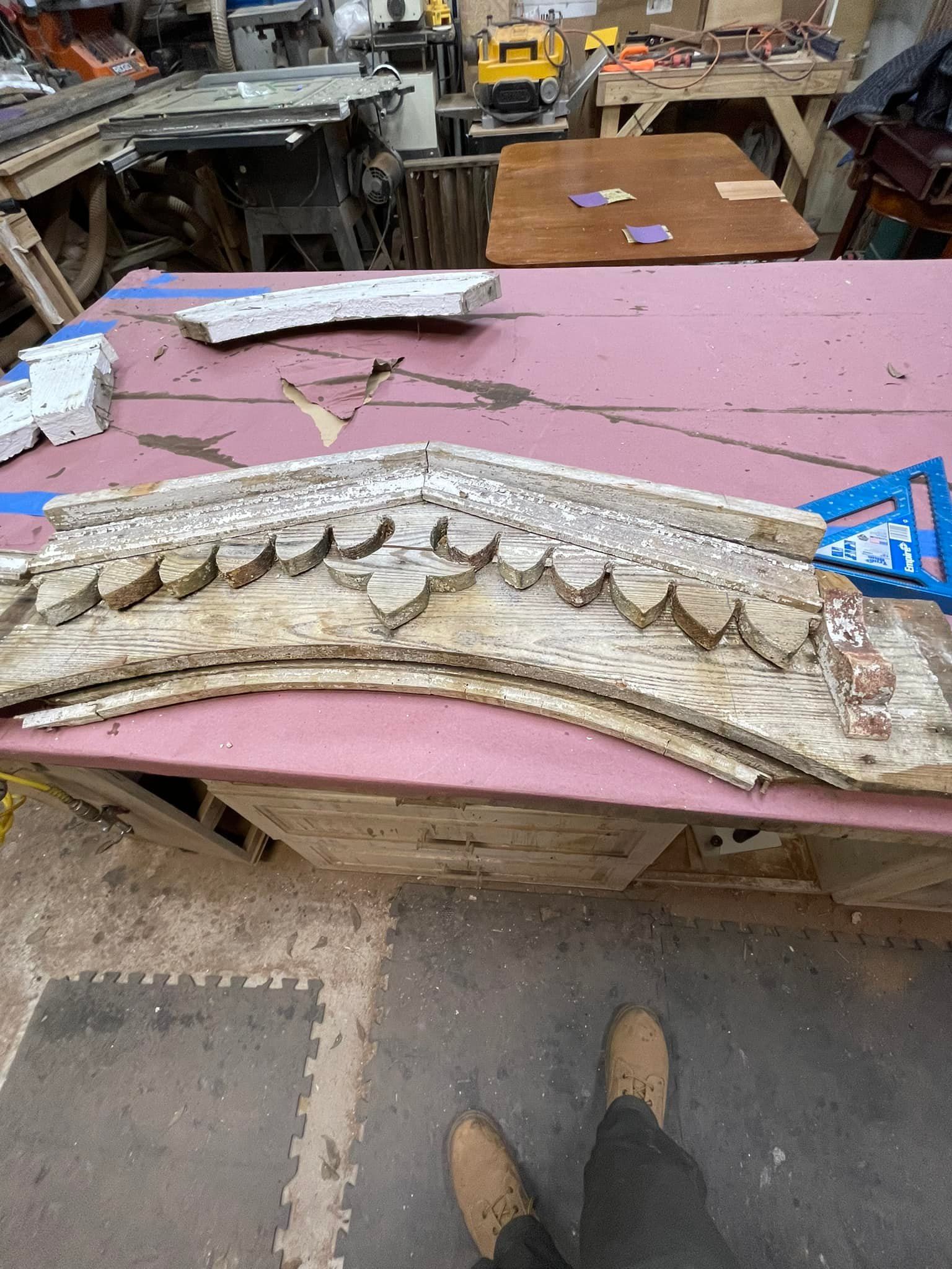 Person's feet near a weathered, ornate wooden architectural piece on a workbench in a workshop.