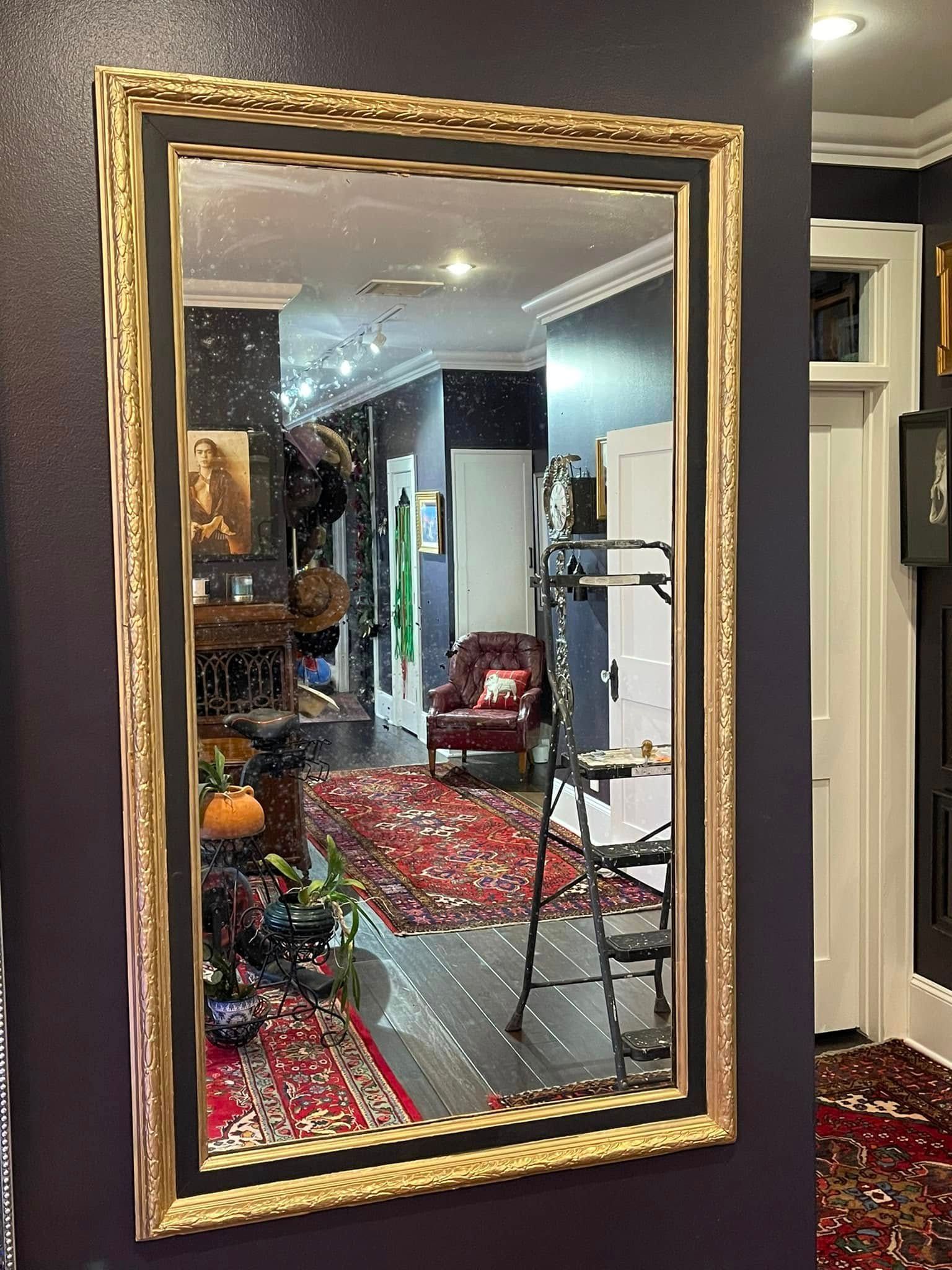 Large rectangular mirror with ornate gold frame, reflecting a room with red rug and furniture.