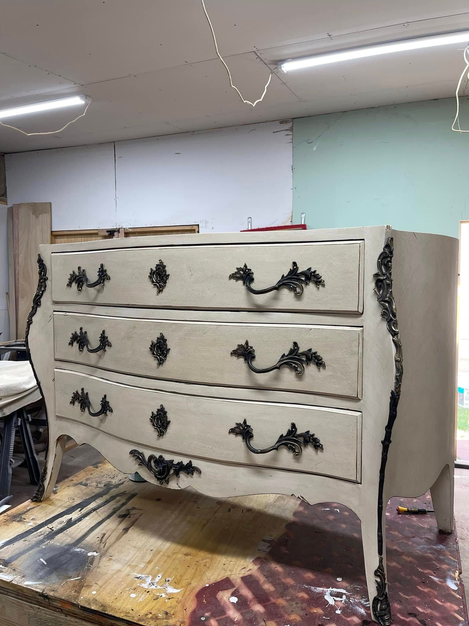Cream-colored, ornate chest of drawers with black hardware, in a workshop setting.