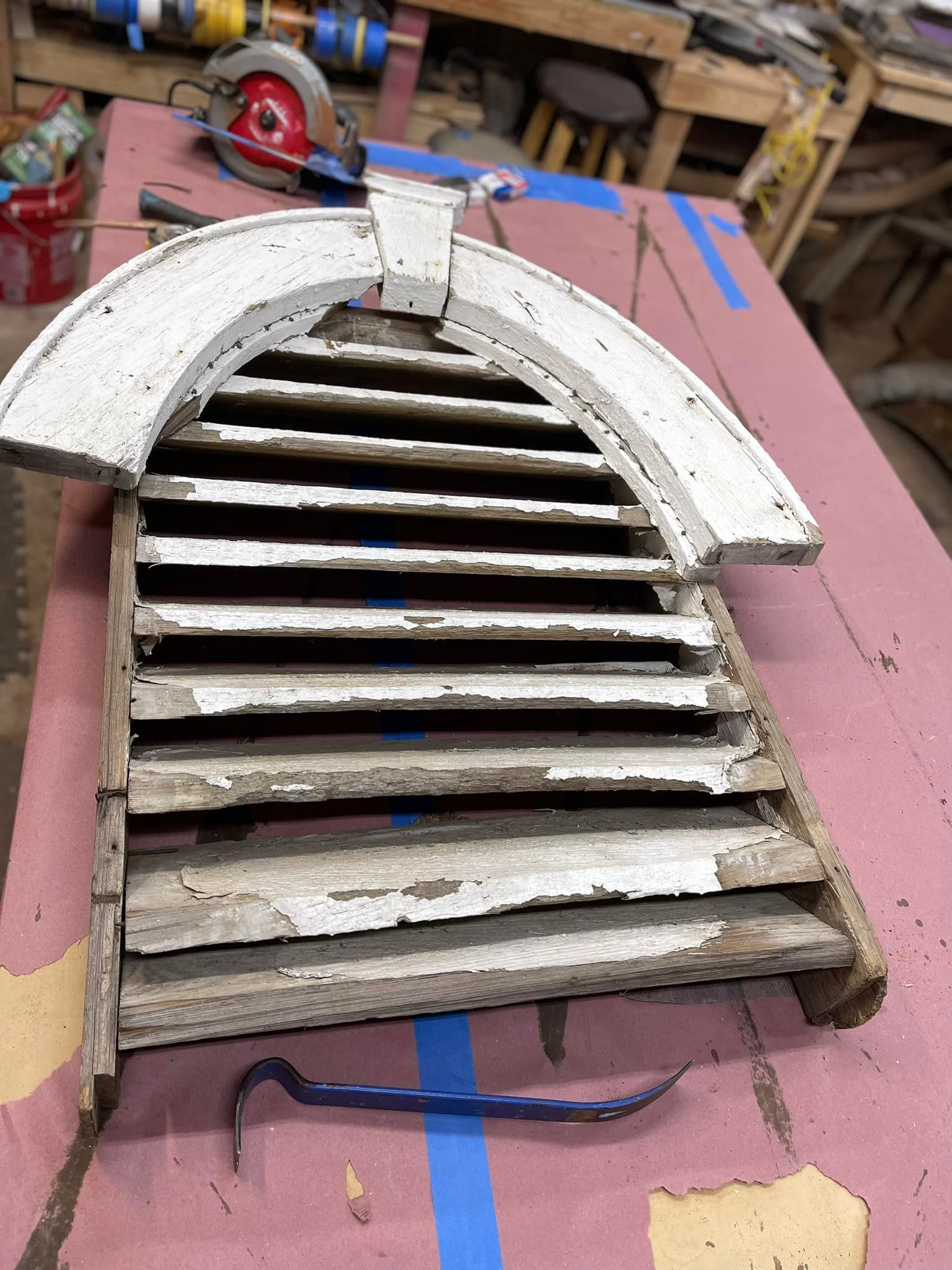 Arched wooden vent with white peeling paint, on a pink surface with a blue tape line and crowbar.