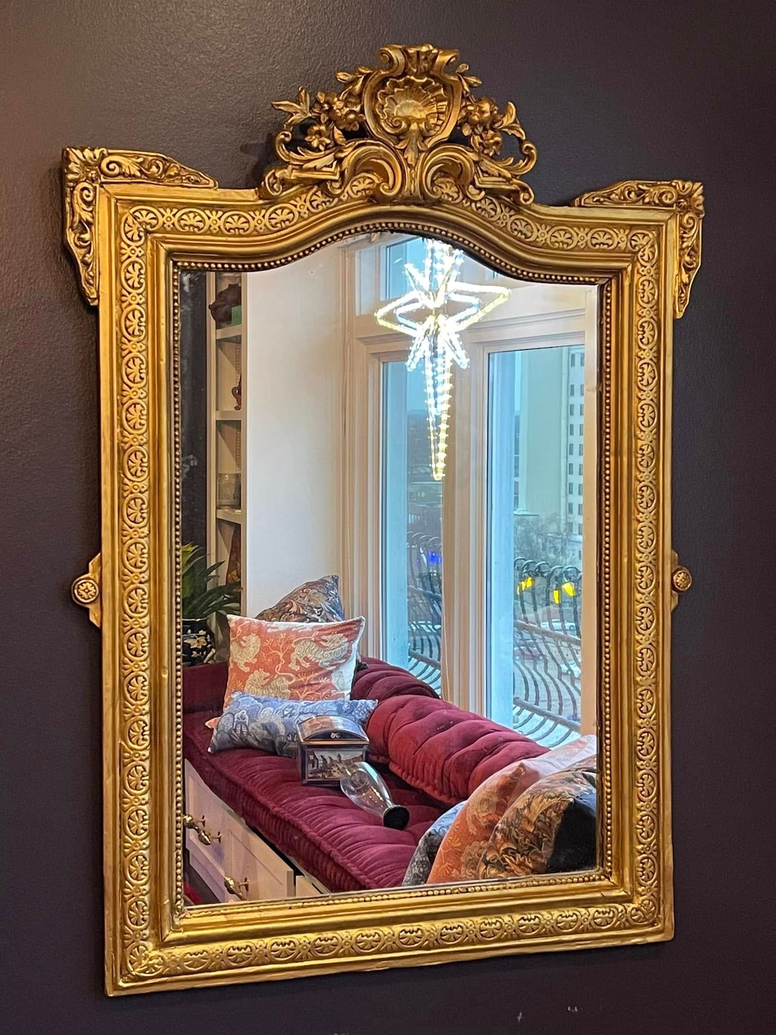 Gold framed mirror reflecting a room with a red cushioned bench, pillows, and a window with a Christmas light.