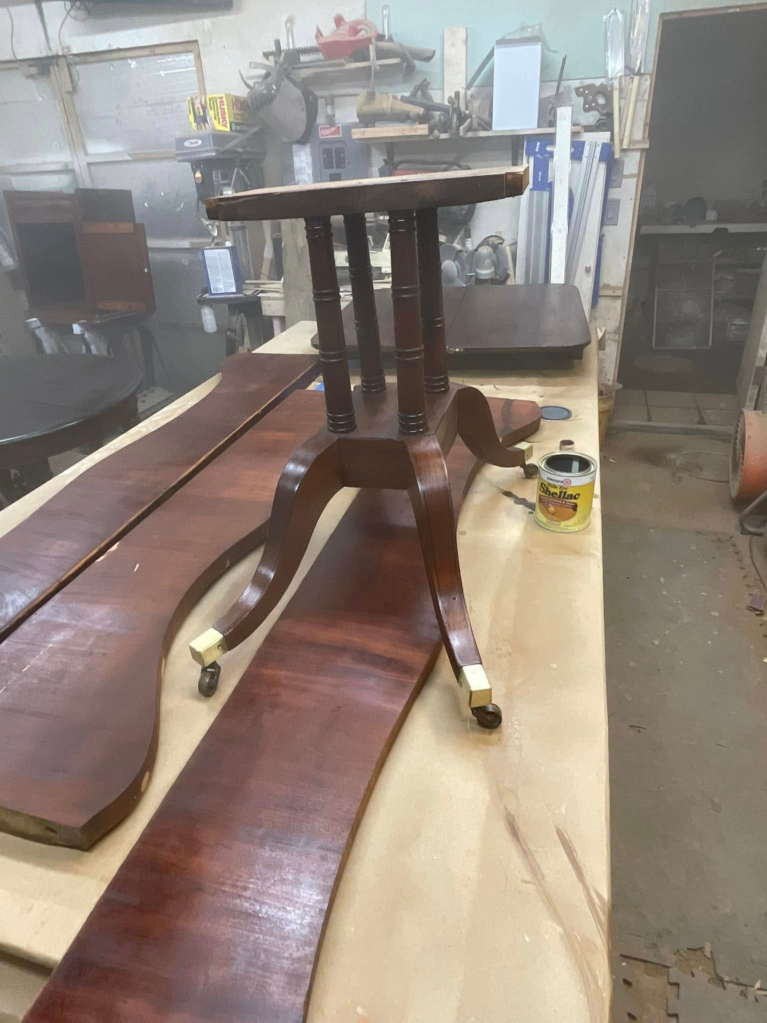 Wooden table with curved legs, sitting on a workbench next to long wood planks, workshop setting.